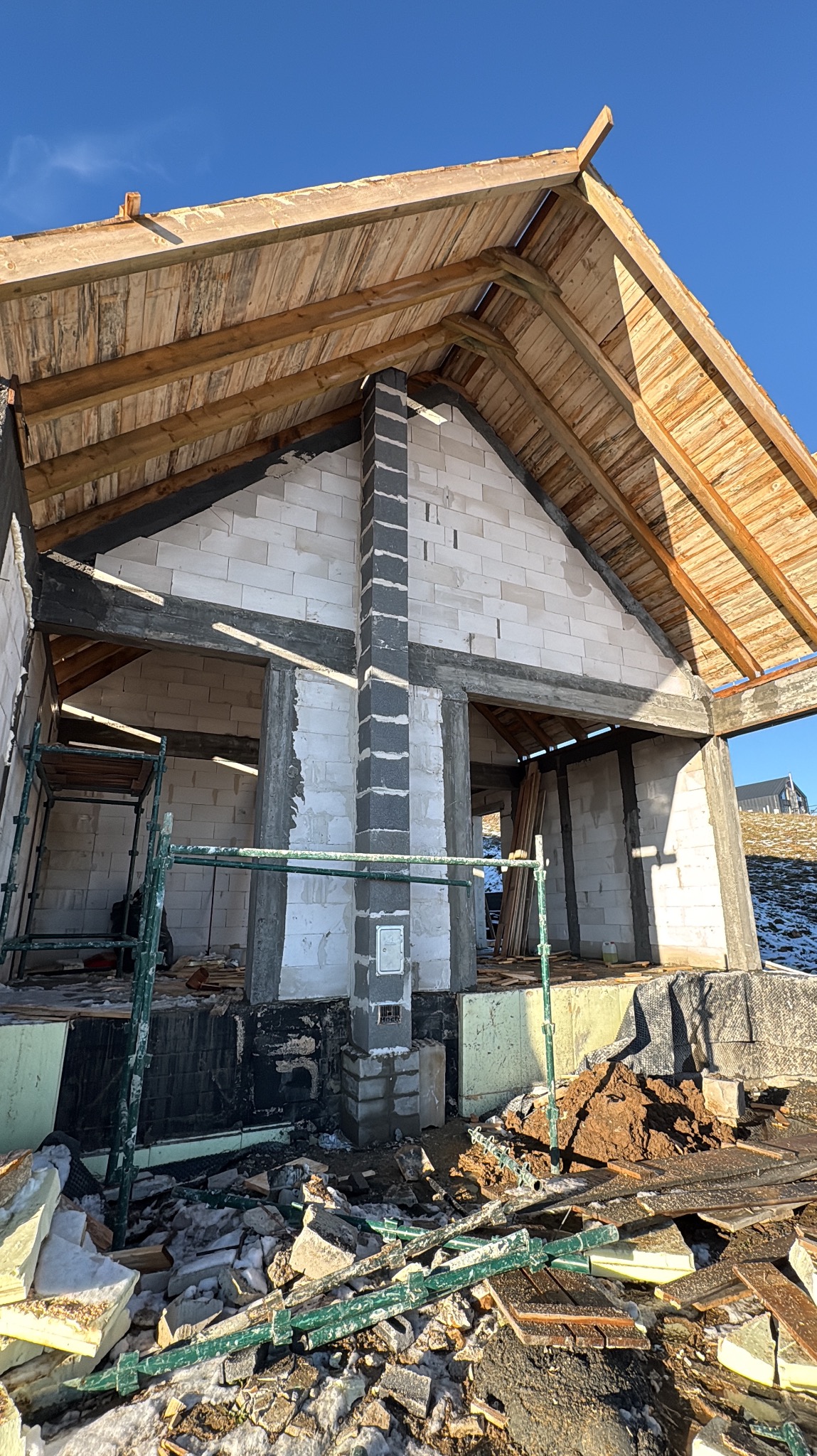 Surowy stan domu z białych bloczków, widoczna więźba dachowa i komin z szarych elementów. Na pierwszym planie gruz i resztki materiałów budowlanych.