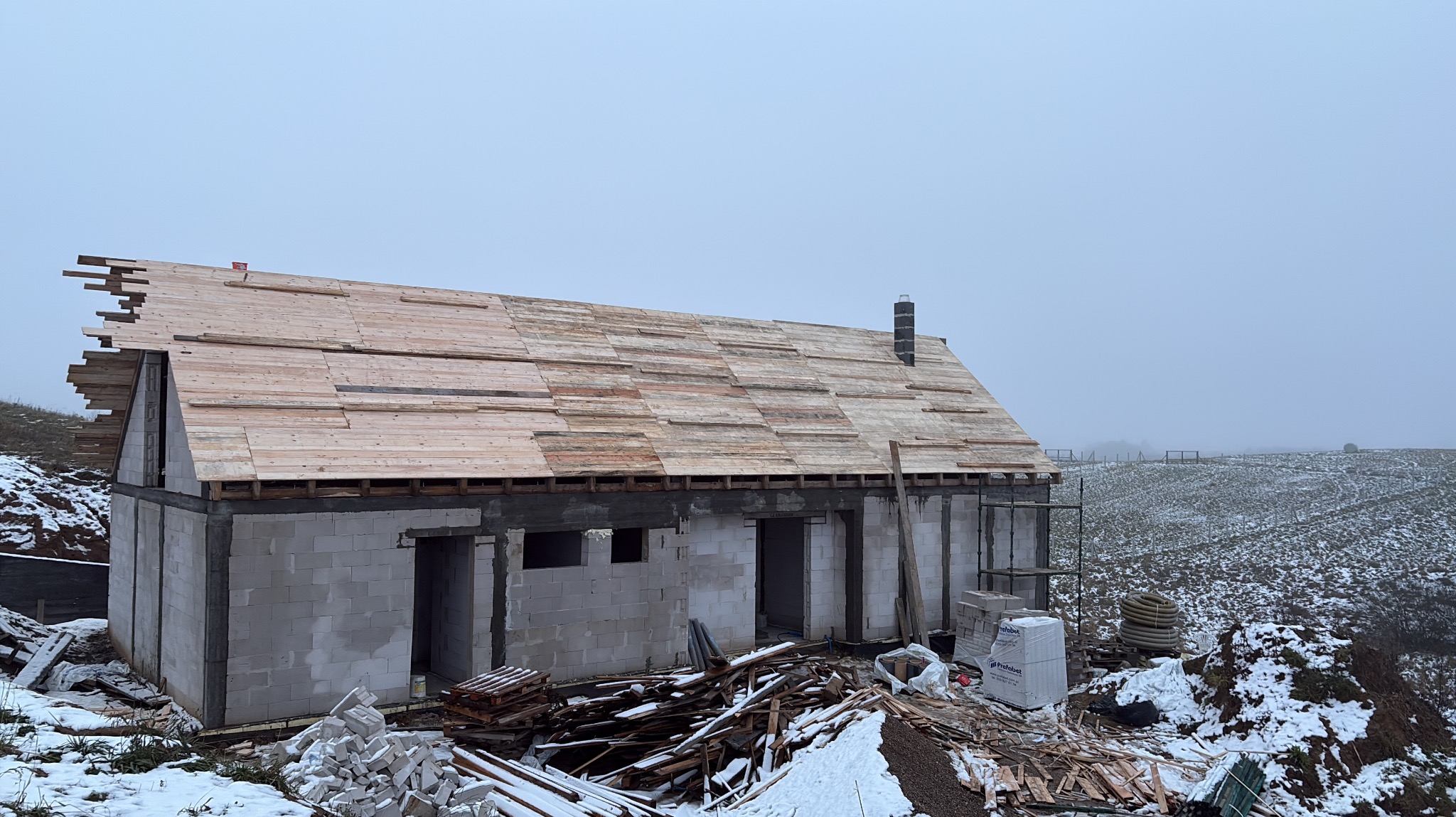 Budowa domu w trakcie: ściany z betonu komórkowego, częściowo ułożona więźba dachowa z desek. Zimowy krajobraz w tle, mgła. Widoczne materiały budowlane.