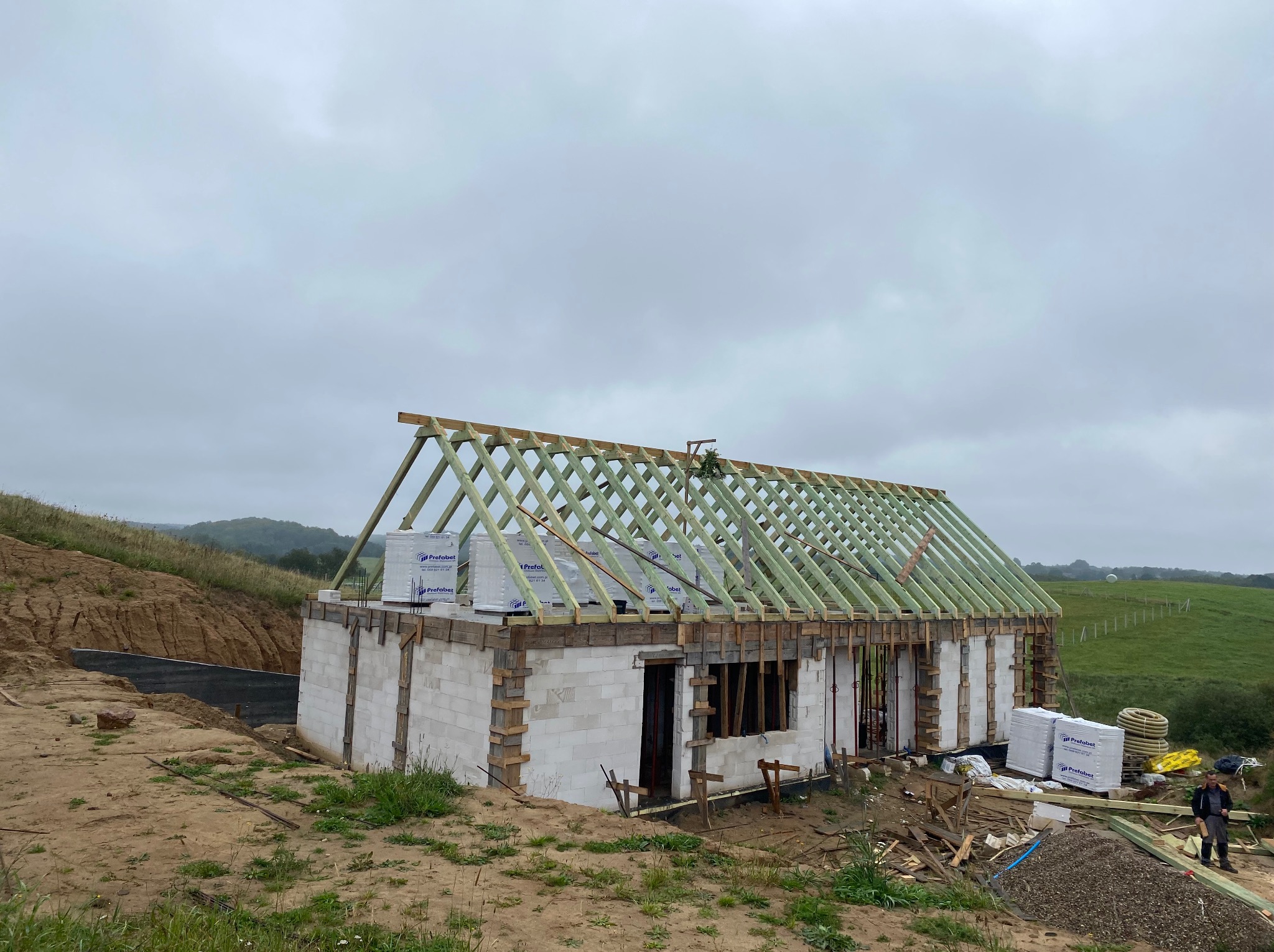 Budowa domu jednorodzinnego w trakcie realizacji: wzniesione ściany z bloczków, konstrukcja więźby dachowej, materiały budowlane na placu. Widok na pagórkowaty teren.
