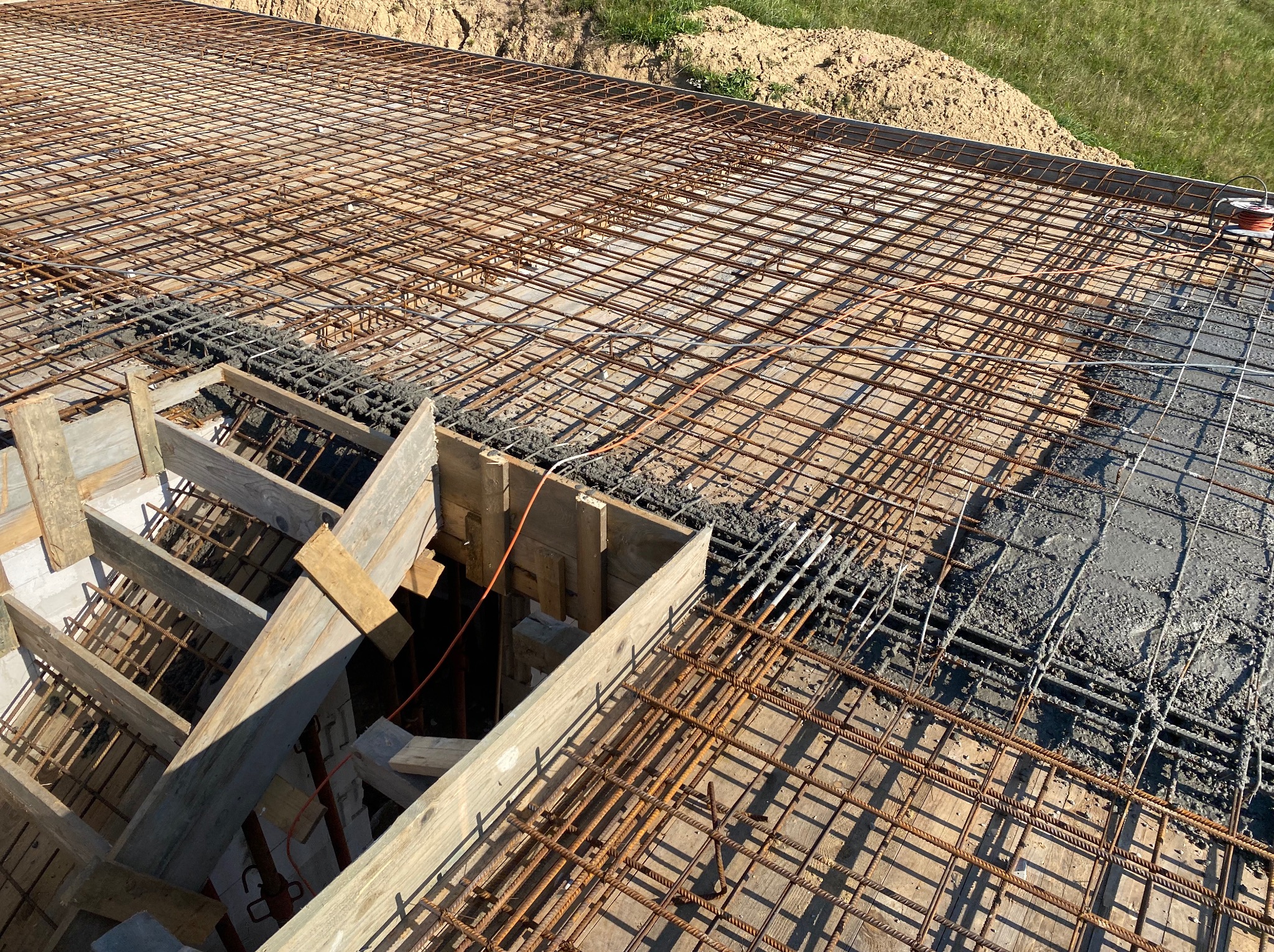 Zbrojenie fundamentów z częściowo wylanym betonem, widoczne drewniane szalunki i siatka zbrojeniowa. Ujęcie z góry, w tle ziemia i trawa.