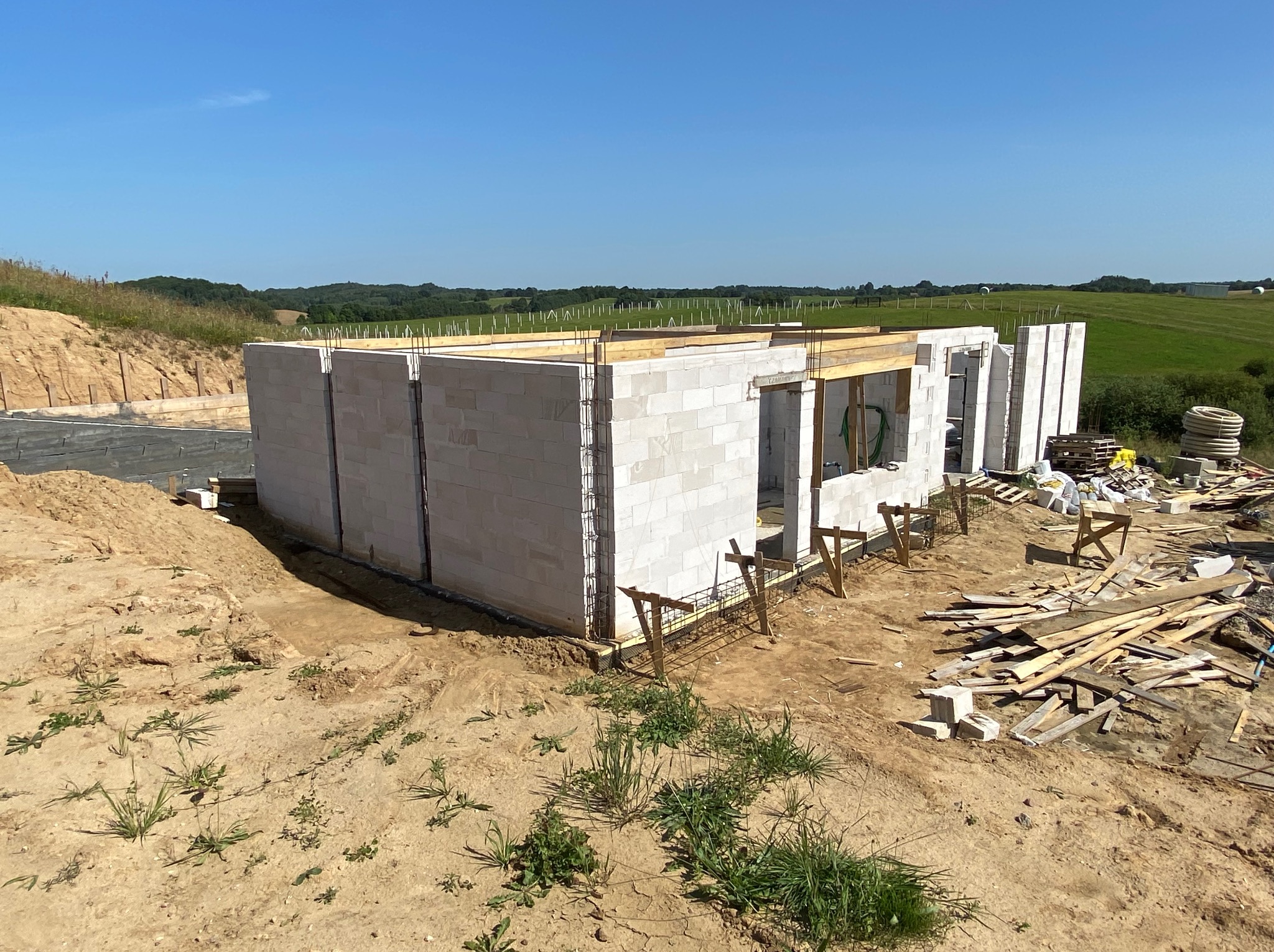 Budowa domu w toku: ściany z betonu komórkowego, zbrojenie, drewniane podpory. W tle zielone pola i błękitne niebo. Widok z poziomu gruntu, w słoneczny dzień.