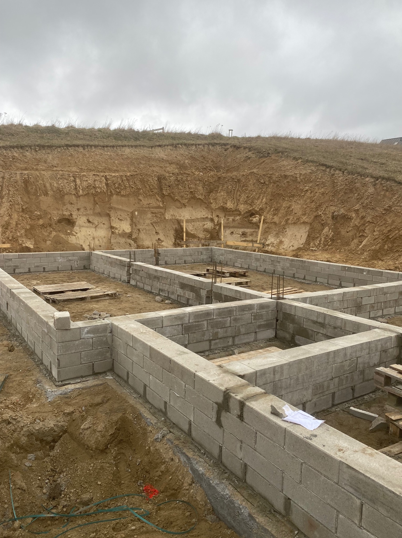 Wysoki kąt na fundamenty z bloczków betonowych, zbrojenie pionowe, wykop ziemny w tle. Biała szmatka leży na murze. Pochmurne niebo.