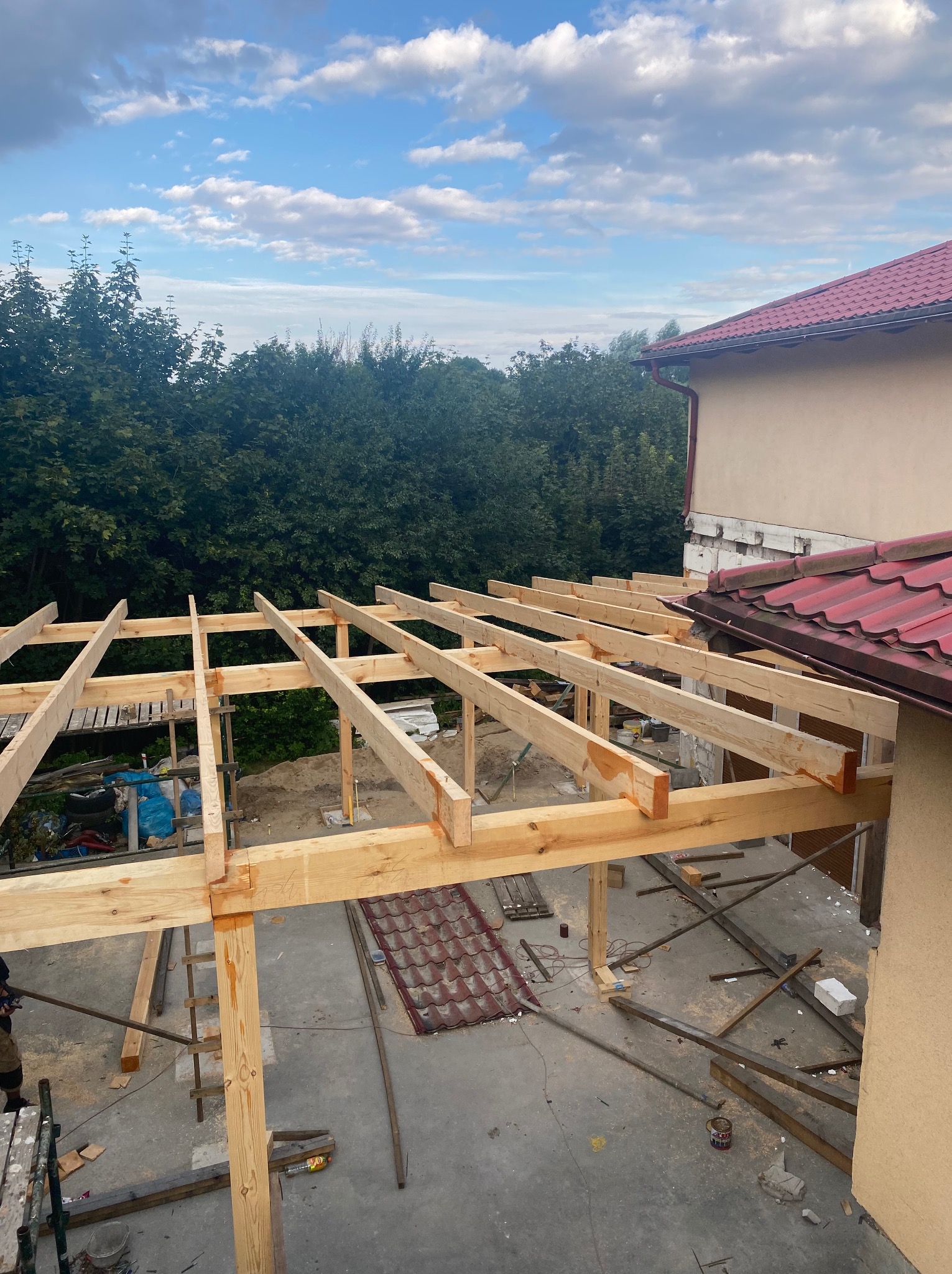 Wznoszenie drewnianej konstrukcji dachu z widocznymi elementami więźby, na tle budynku z czerwoną dachówką i zieleni drzew, w słoneczny dzień.