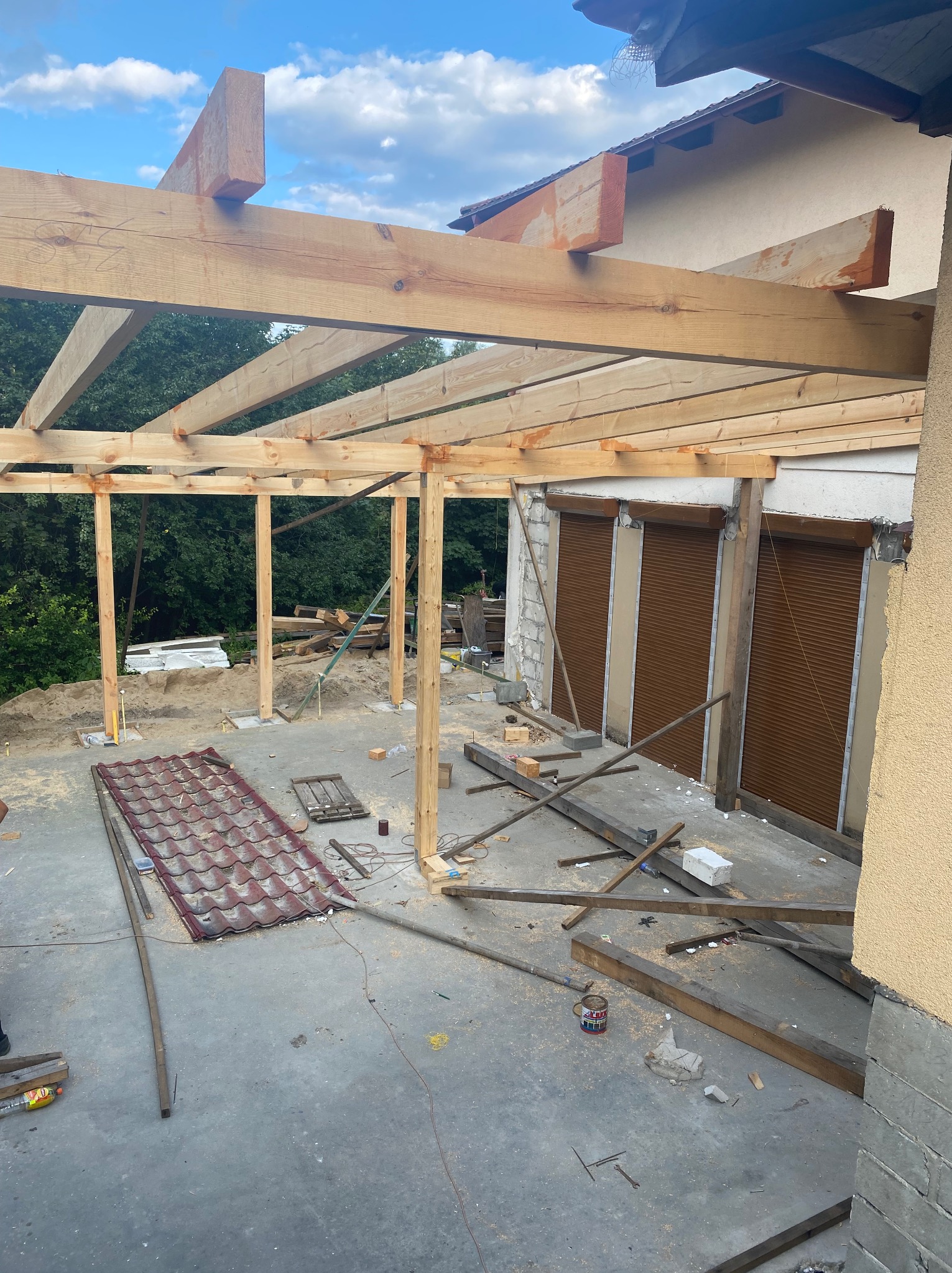 Wznoszenie drewnianej konstrukcji z belek na betonowej powierzchni, widoczne elementy dachu i rolety okienne. Prace budowlane w toku, narzędzia i materiały budowlane w tle.