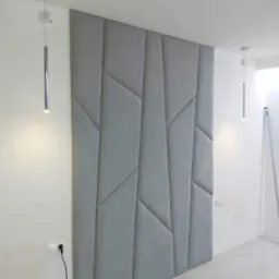 Szary panel tapicerowany na ścianie z geometrycznym wzorem, oświetlony przez dwie chromowane lampy wiszące, widoczne gniazdka elektryczne i ślady po pracach wykończeniowych.