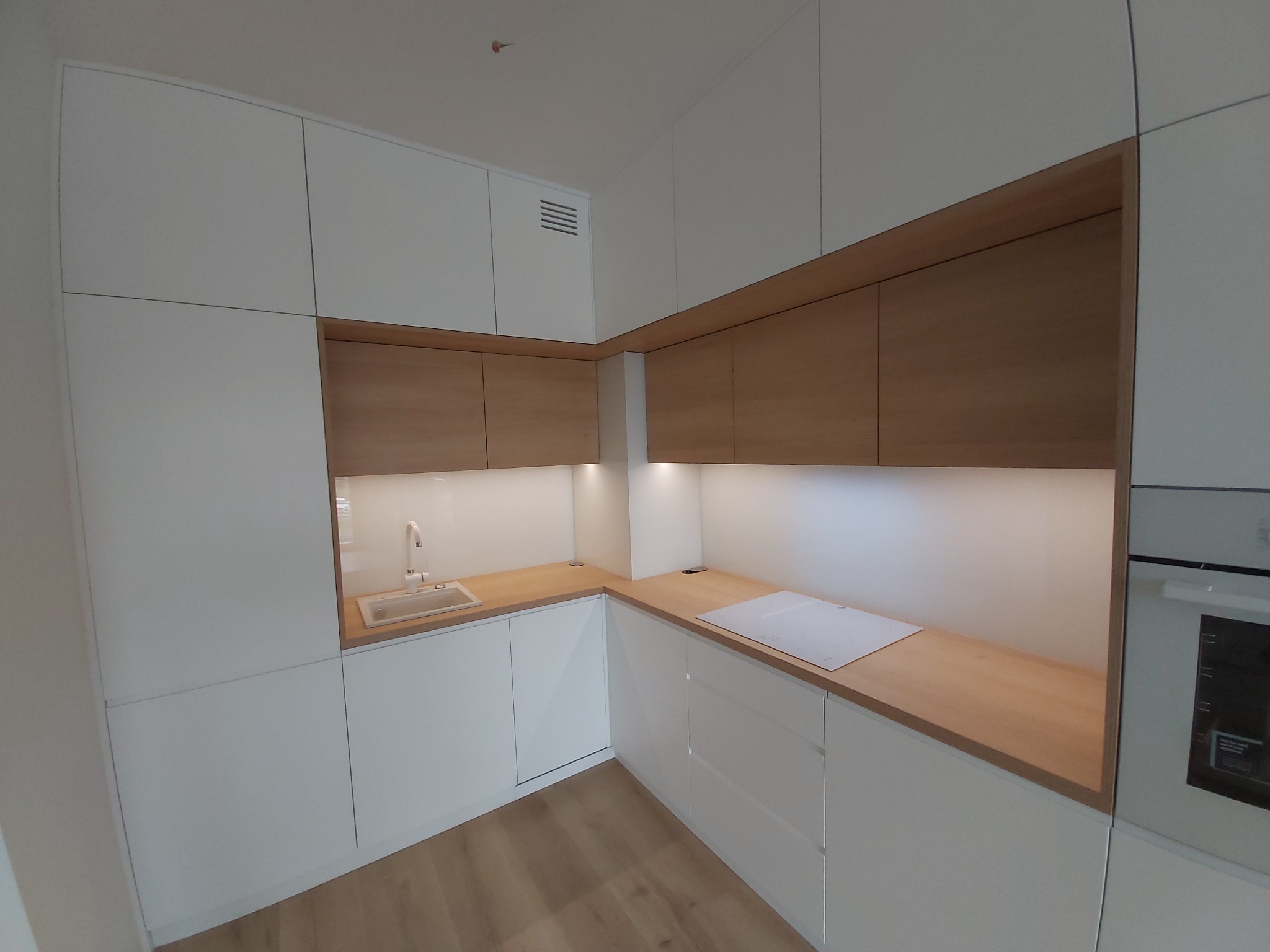 Nowoczesna, narożna kuchnia z białymi, lakierowanymi frontami i blatem z jasnego drewna, oświetlona punktowo, z minimalistyczną armaturą i płytą indukcyjną.