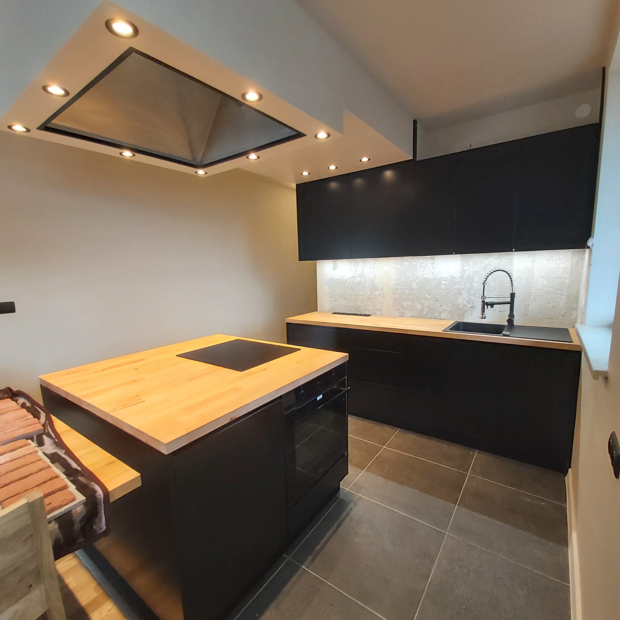 Nowoczesna kuchnia z czarnymi szafkami i blatem z jasnego drewna, wyspa kuchenna z płytą indukcyjną i piekarnikiem, szara podłoga i oświetlenie punktowe w suficie