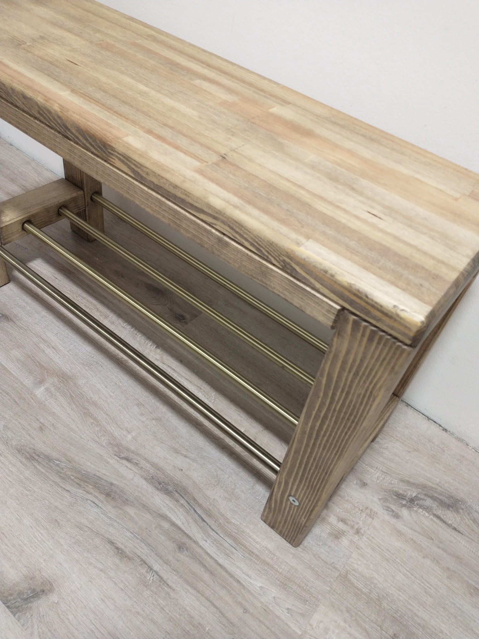 Drewniana ławka z trzema metalowymi poprzeczkami na buty, stojąca na podłodze imitującej drewno, przy białej ścianie.