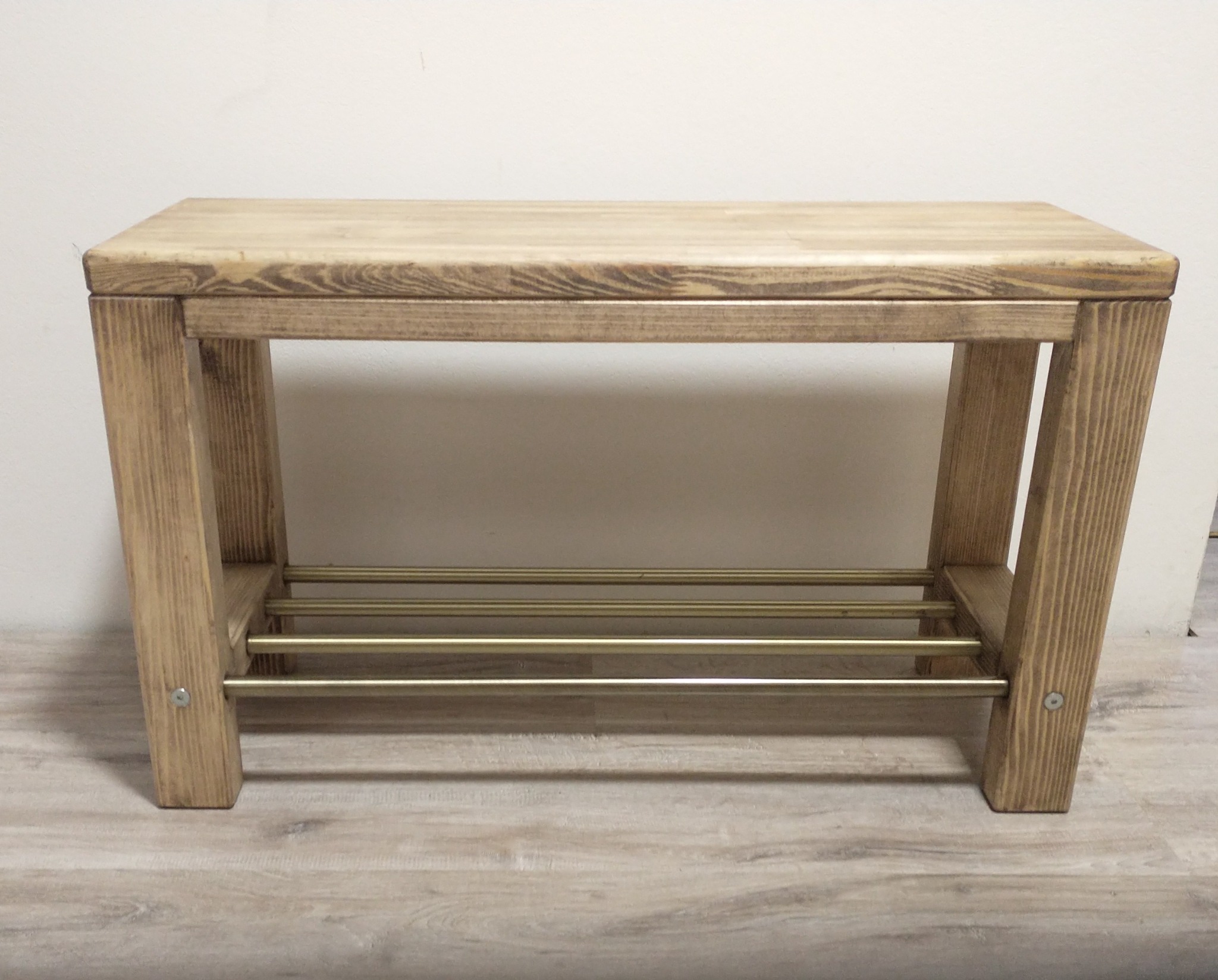 Drewniana ławka z brązowionego drewna, z trzema złotymi poprzeczkami na buty, ustawiona na panelach podłogowych.