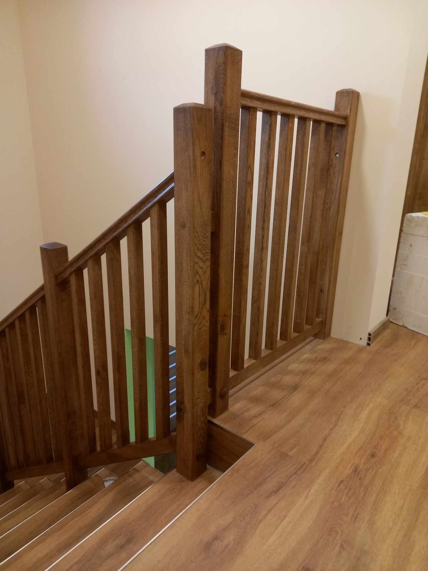 Drewniane schody z balustradą o ciepłym odcieniu, widoczne z góry, z fragmentem ściany w tle i podłogą imitującą drewno