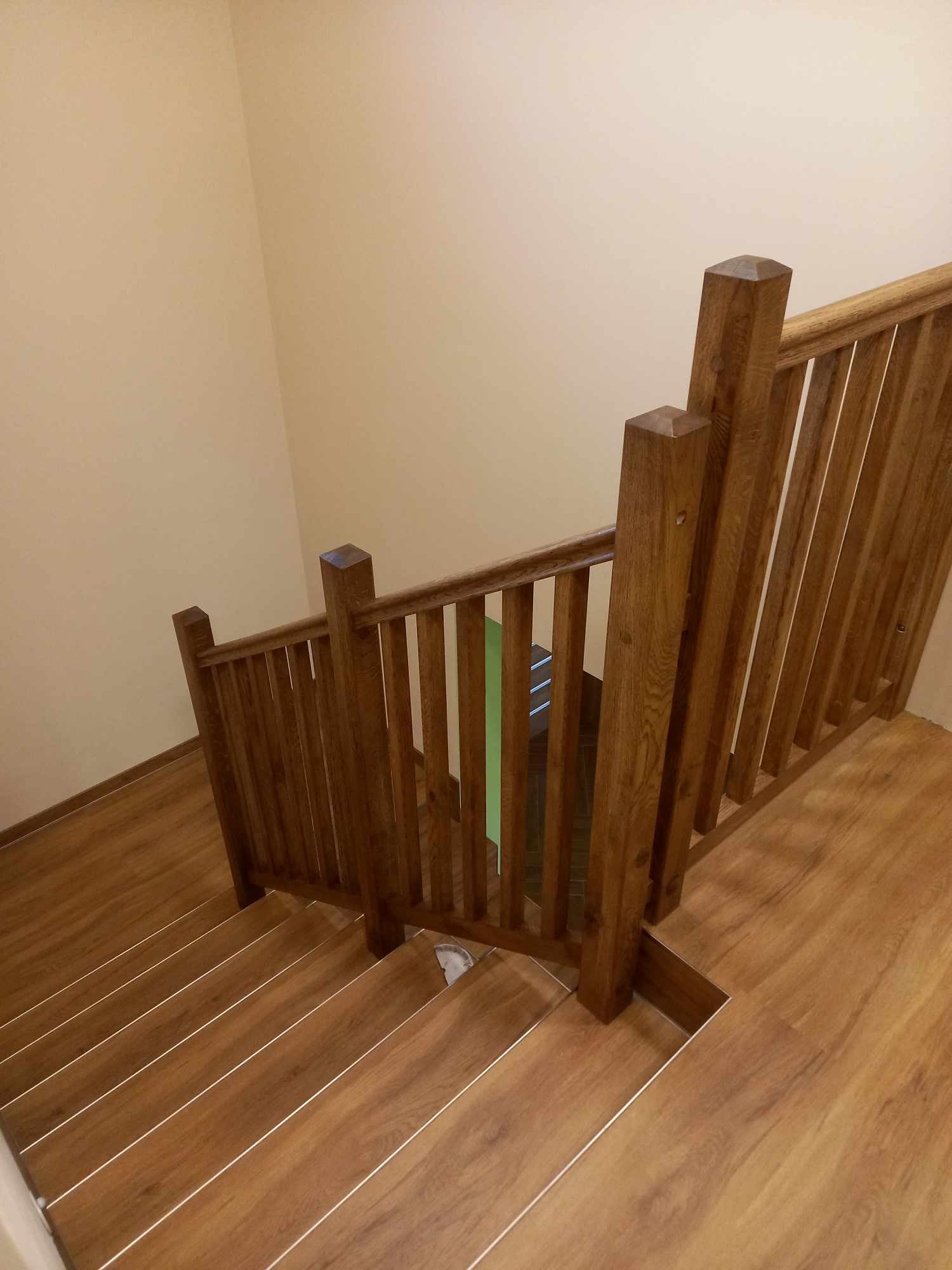 Klatka schodowa z drewnianymi schodami i balustradą w ciepłym odcieniu brązu, widok z góry, ściana pomalowana na beżowo.