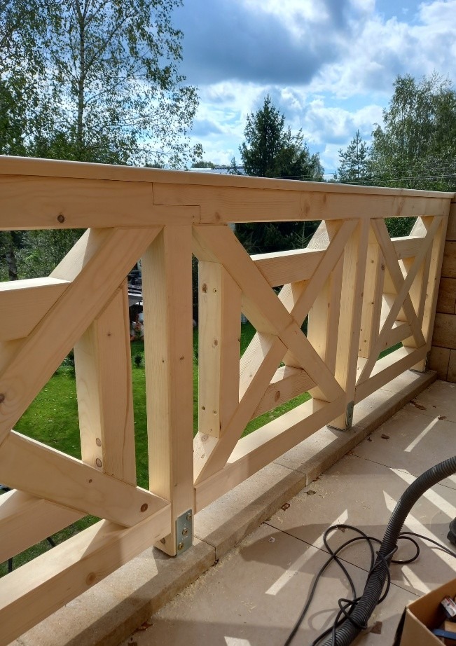 Surowe drewniane balustrady balkonowe z krzyżowym wzorem, widok na zielony ogród i błękitne niebo z chmurami, w tle widoczny odkurzacz przemysłowy i karton z narzędziami.