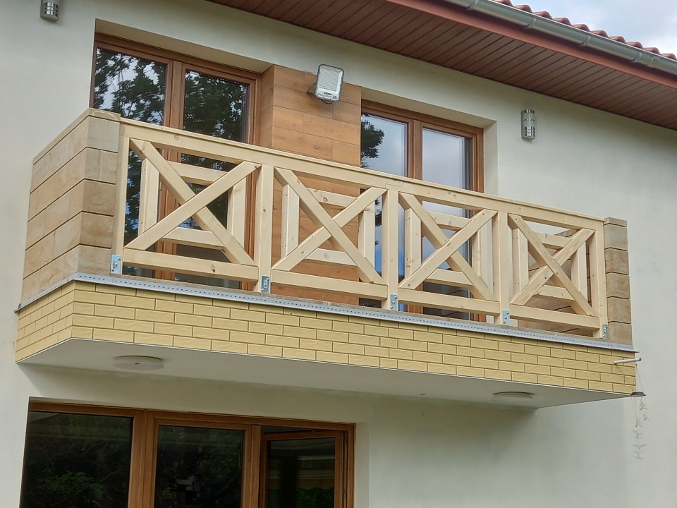 Wykończony balkon z drewnianą balustradą w kształcie krzyży, obłożony płytkami imitującymi cegłę w dwóch odcieniach, widok z dołu na elewację budynku.