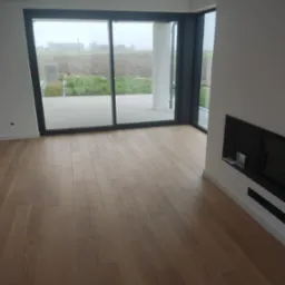 Jasne panele podłogowe z widocznym wzorem słoj&oacute;w, ułożone w nowoczesnym wnętrzu z dużym oknem przesuwnym i minimalistycznym kominkiem.