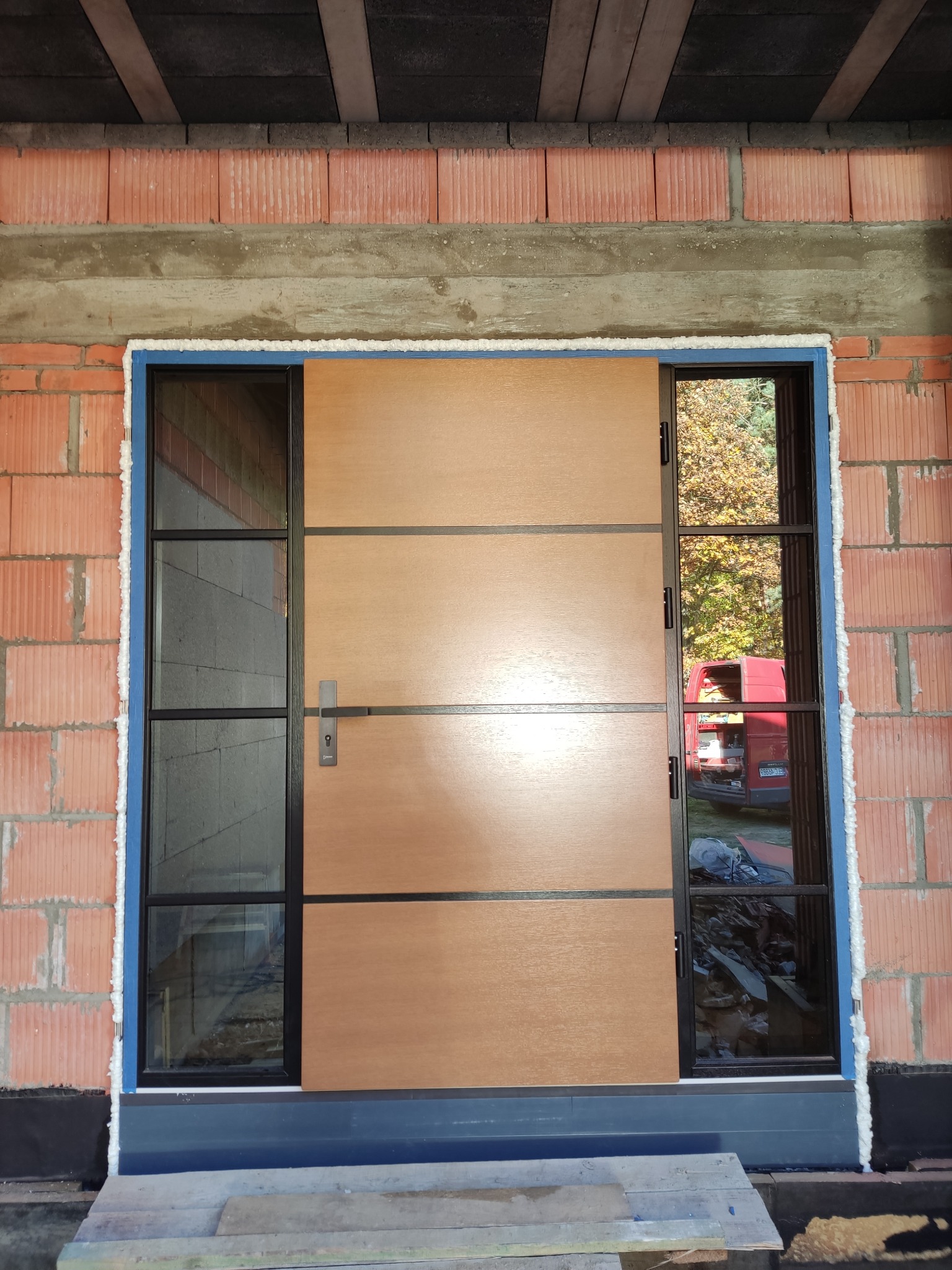 Brązowe drzwi wejściowe z czarną ramą i bocznymi przeszkleniami, zamontowane w surowym murze z czerwonej cegły, widoczna piana montażowa i zaparkowany czerwony samochód w tle.