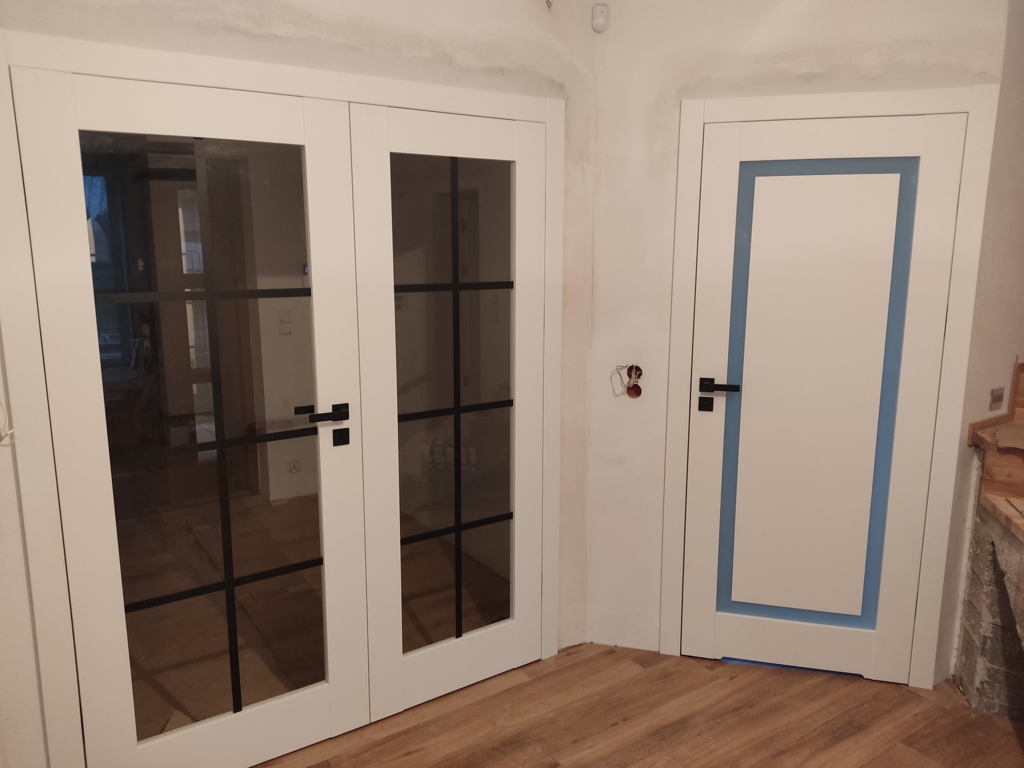 Dwa rodzaje białych drzwi wewnętrznych z czarnymi klamkami: podwójne, przeszklone z czarnymi szprosami i pojedyncze z niebieską wstawką, zamontowane w nowym budynku z drewnianą podłogą.