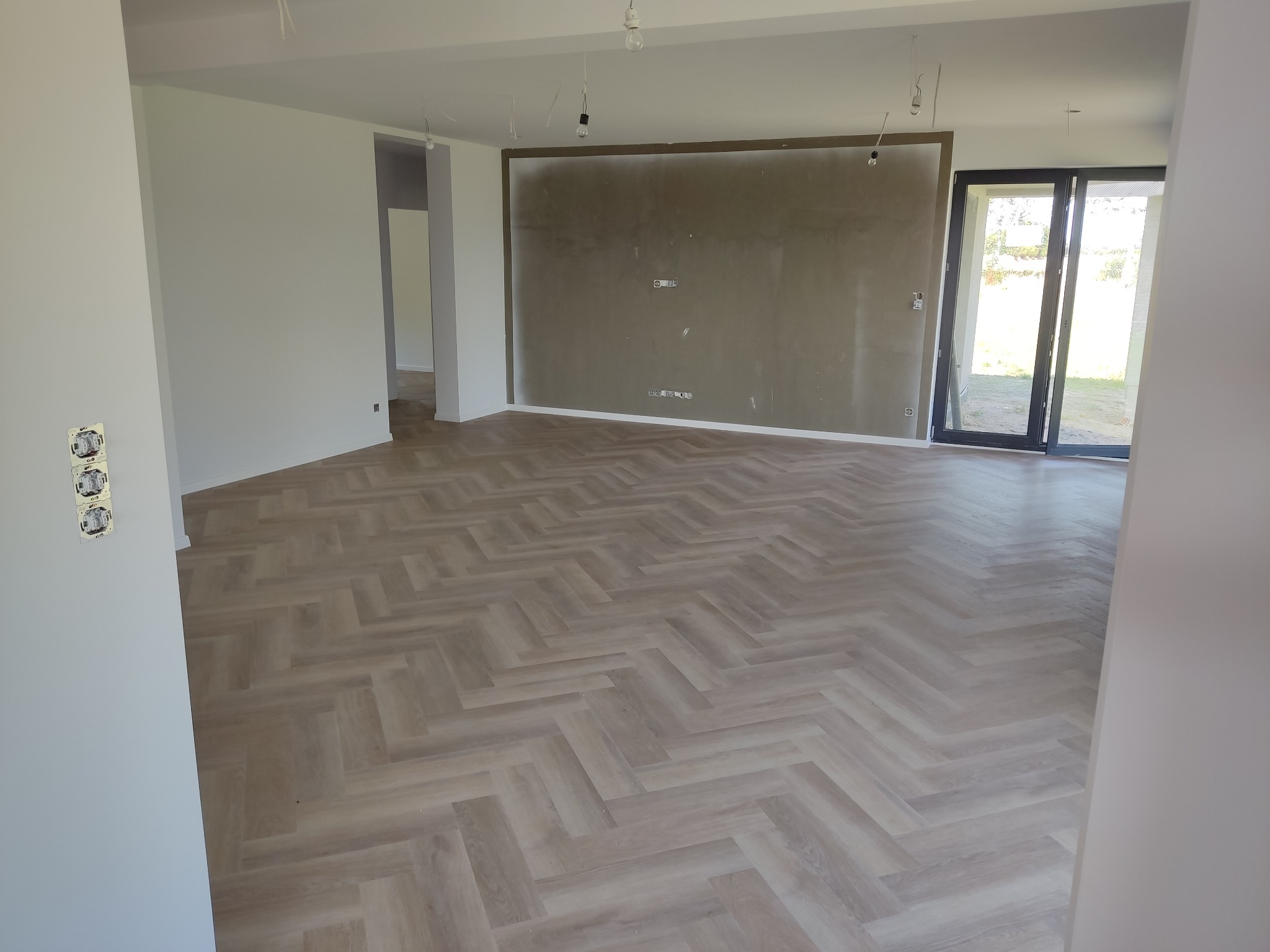 Jasne panele ułożone we wzór jodełki w przestronnym, surowym wnętrzu z widocznymi puszkami elektrycznymi i niedokończonymi ścianami.