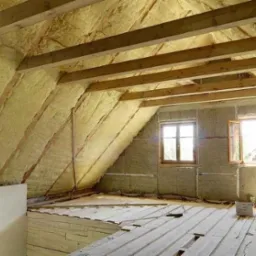 Poddasze z ociepleniem z wełny mineralnej, widoczne drewniane belki stropowe i częściowo ułożona podłoga z desek, dwa okna dachowe z otwartymi skrzydłami.