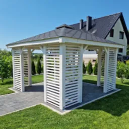 Biała, drewniana altana z ażurowymi ścianami na tle zadbanego trawnika i domu z ciemnym dachem, widok z zewnątrz, słoneczny dzień.