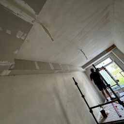 Monte Lux Adrian Wensierski - Praca na wysokości: mężczyzna na rusztowaniu szpachluje sufit w skosie, widoczne płyty karton-gips i narzędzia. Okno z widokiem na zieleń w tle.