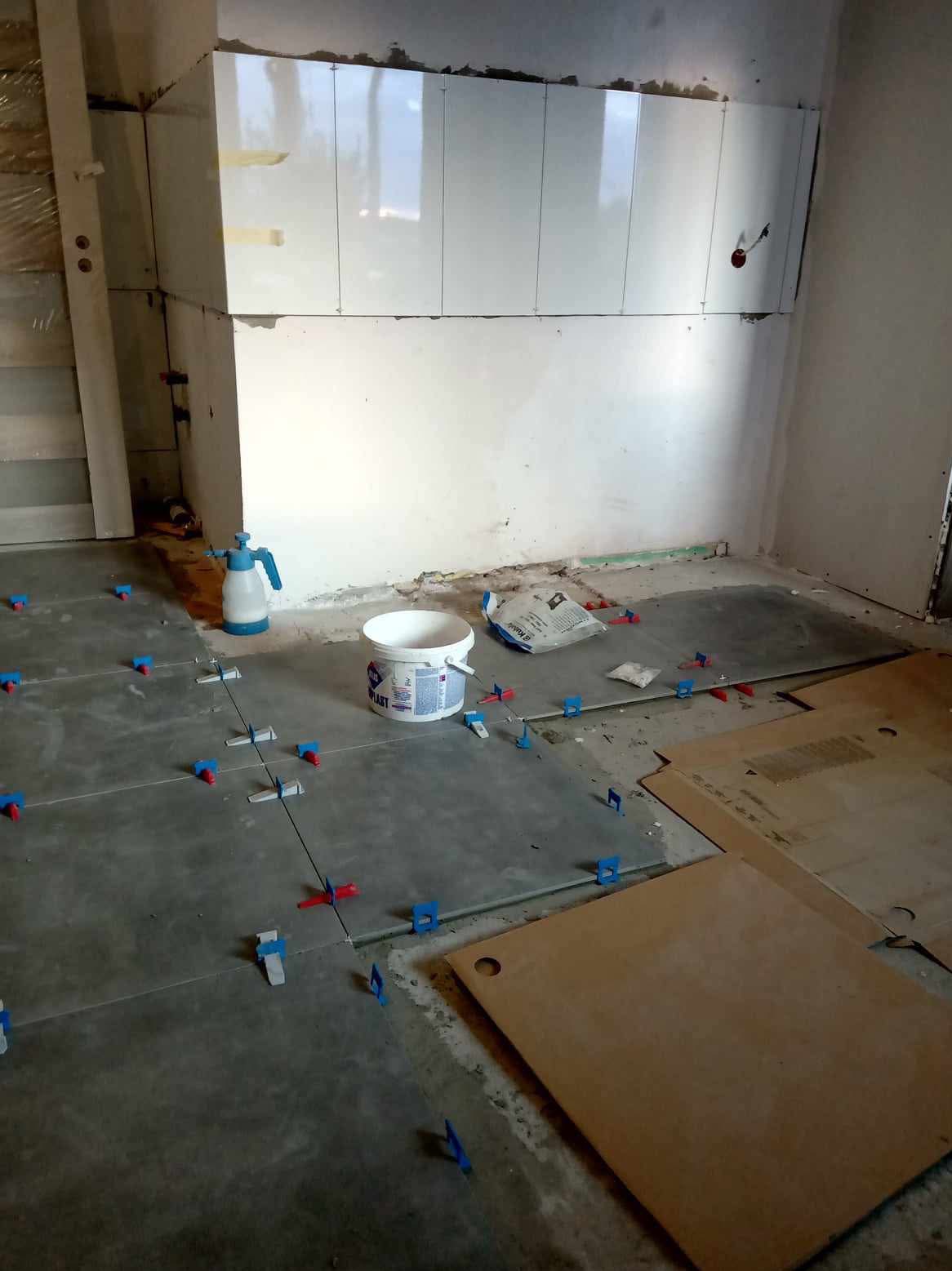 Układanie szarych płytek podłogowych w trakcie remontu, z widocznymi krzyżykami dystansowymi, wiaderkiem z klejem i częściowo wyłożonymi płytkami ściennymi w tle.