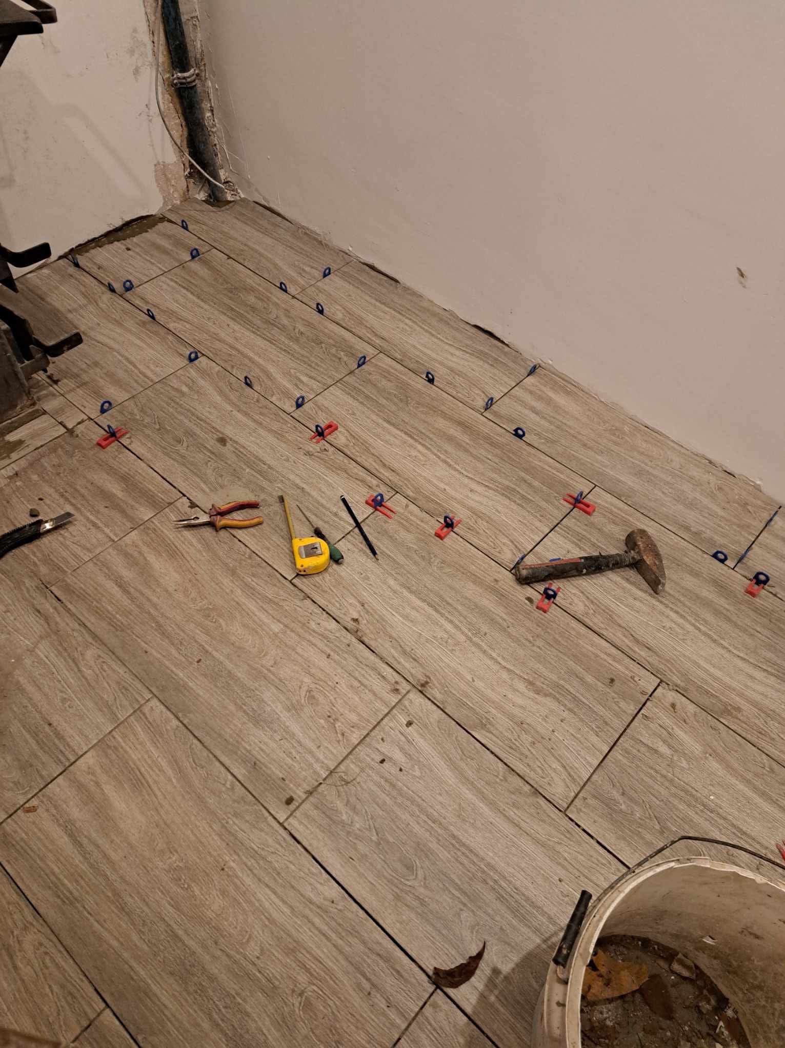 Układanie płytek podłogowych imitujących drewno z użyciem poziomujących klipsów i klinów, widoczne narzędzia: młotek, miarka, kombinerki i ołówek.