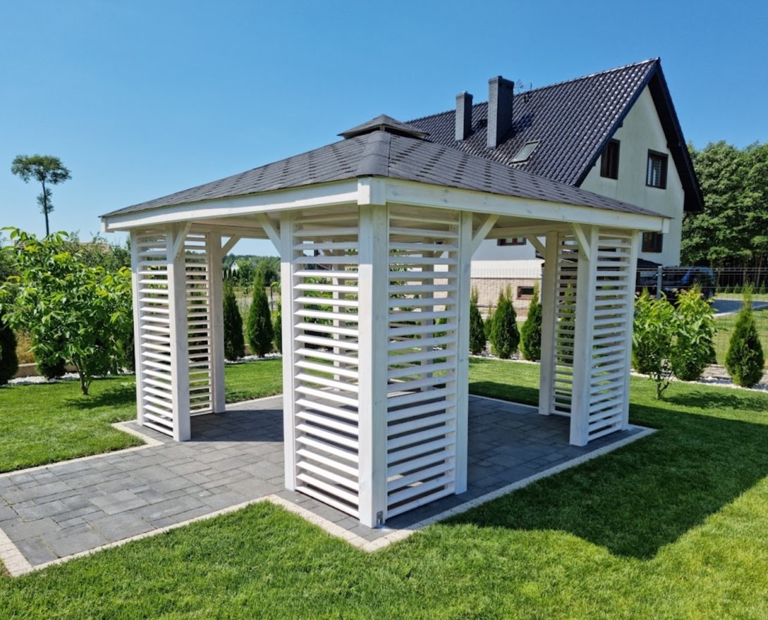 Biała, drewniana altana z ażurowymi ścianami na tle zadbanego trawnika i domu z ciemnym dachem, widok z zewnątrz, słoneczny dzień.