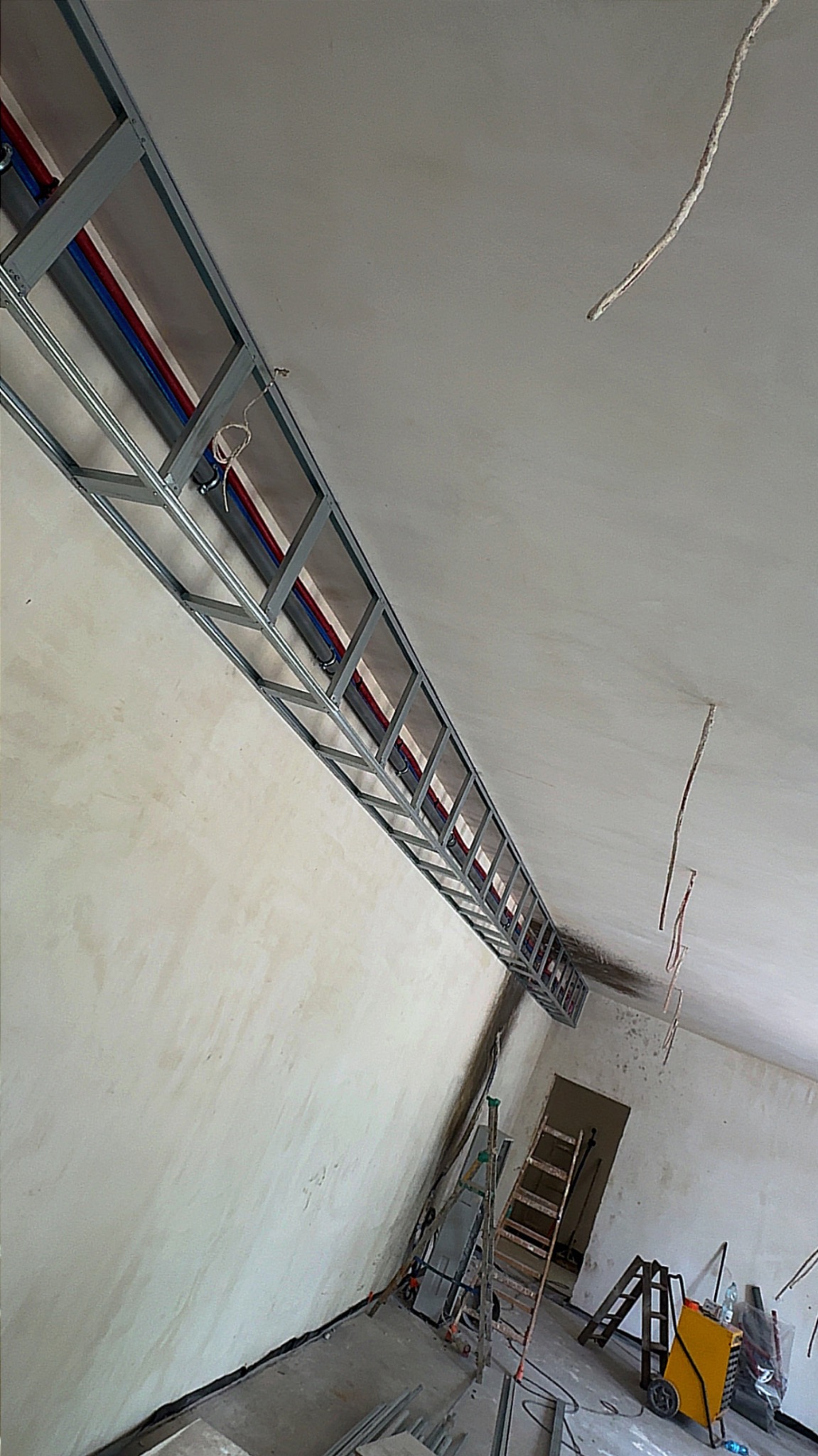 Montaż metalowej konstrukcji pod sufitem z widocznymi instalacjami (rury, przewody). W tle drabiny i narzędzia w surowym wnętrzu.