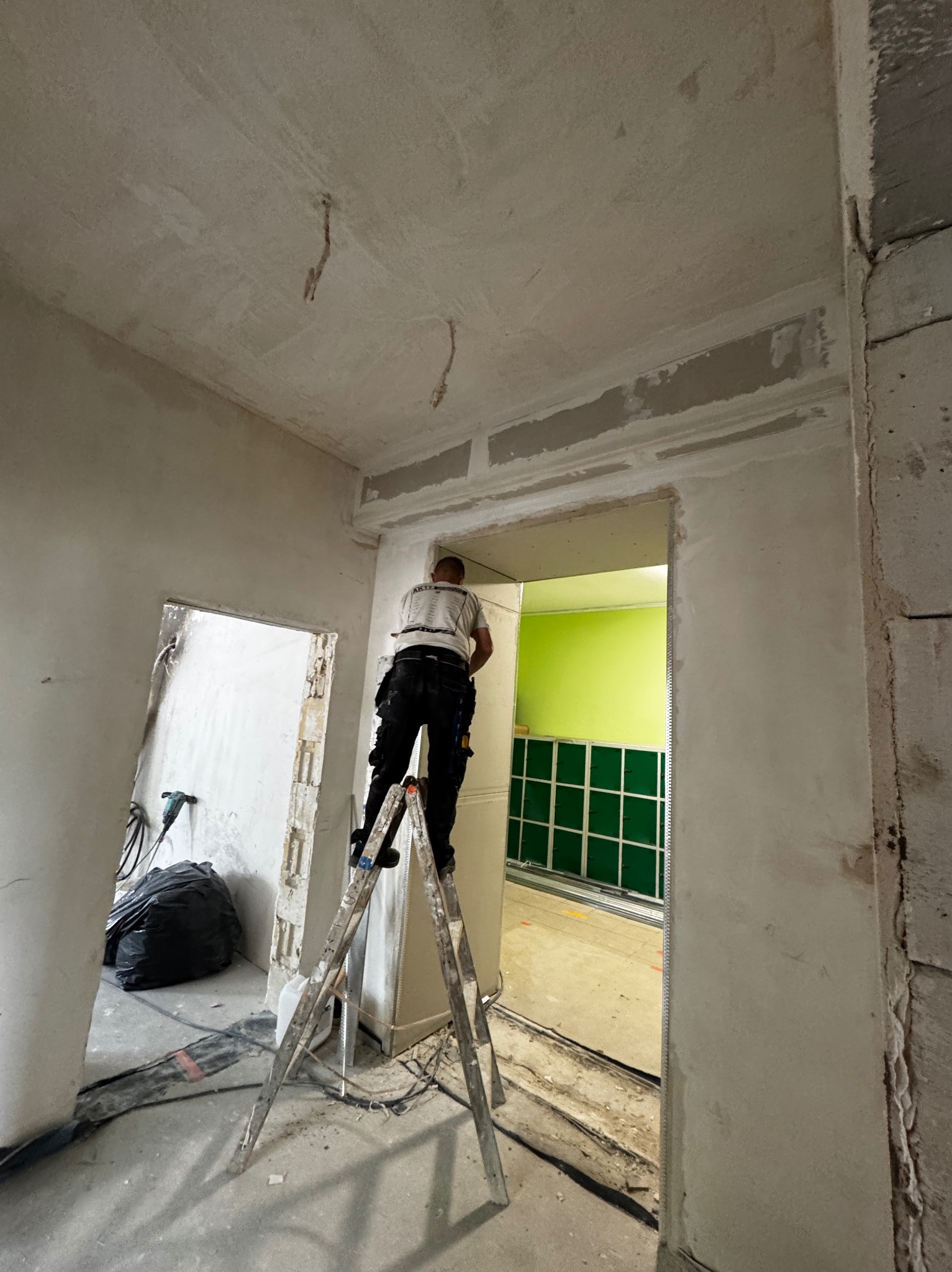 Prace wykończeniowe w Mydlitach: mężczyzna na drabinie montuje płytę karton-gips w futrynie drzwiowej. Widoczne surowe ściany i narzędzia.