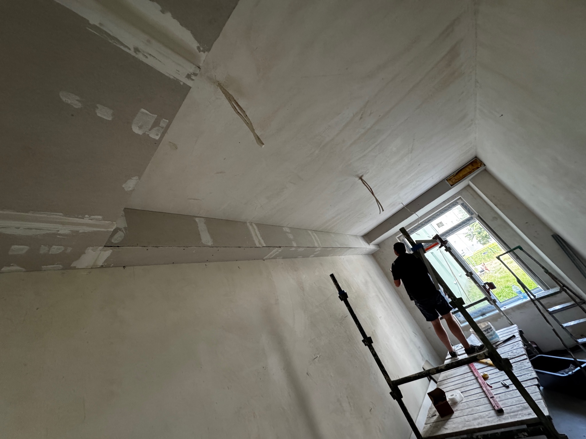 Praca na wysokości: mężczyzna na rusztowaniu szpachluje sufit w skosie, widoczne płyty karton-gips i narzędzia. Okno z widokiem na zieleń w tle.