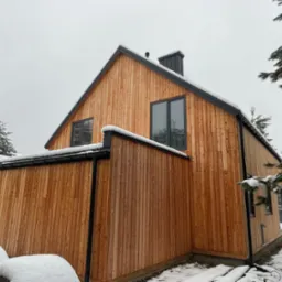 Nowoczesny dom z drewnianą elewacją w zimowej scenerii, widoczne śnieżne czapy na dachu i w ogrodzie, pionowe deski elewacyjne o ciepłym odcieniu drewna, czarne obramowania okien i rynny.