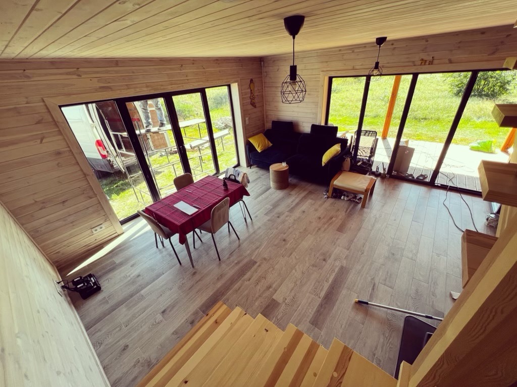 Wnętrze domu z drewnianą boazerią, dużymi oknami z widokiem na zewnątrz, częściowo widocznymi schodami drewnianymi, czarną sofą z żółtymi poduszkami, stołem z czerwonym obrusem i wiszącymi lampami...