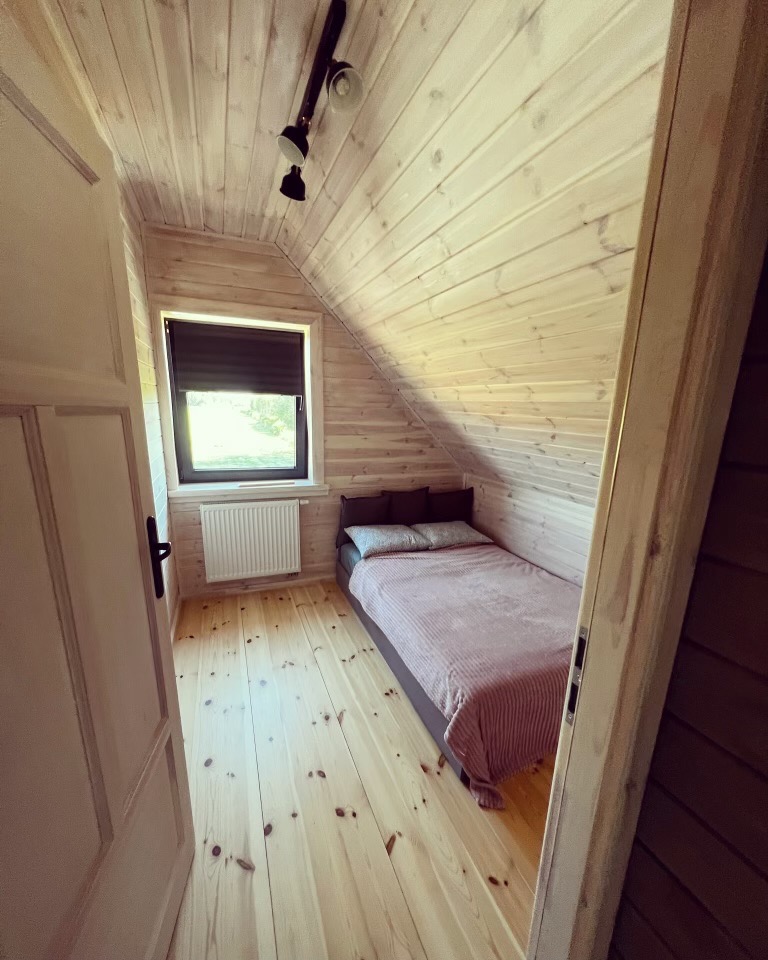 Pokój na poddaszu wykończony boazerią, widoczne okno z roletą, łóżko z narzutą w odcieniach różu i szarości, oraz lampa sufitowa na szynie.