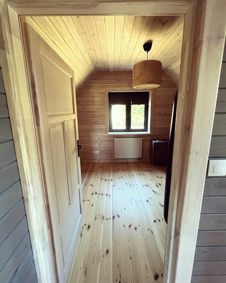 Wnętrze poddasza z drewnianą boazerią, widok przez otwarte drzwi, jasna podłoga z widocznymi sękami, okno z grzejnikiem pod spodem i pleciona lampa wisząca.