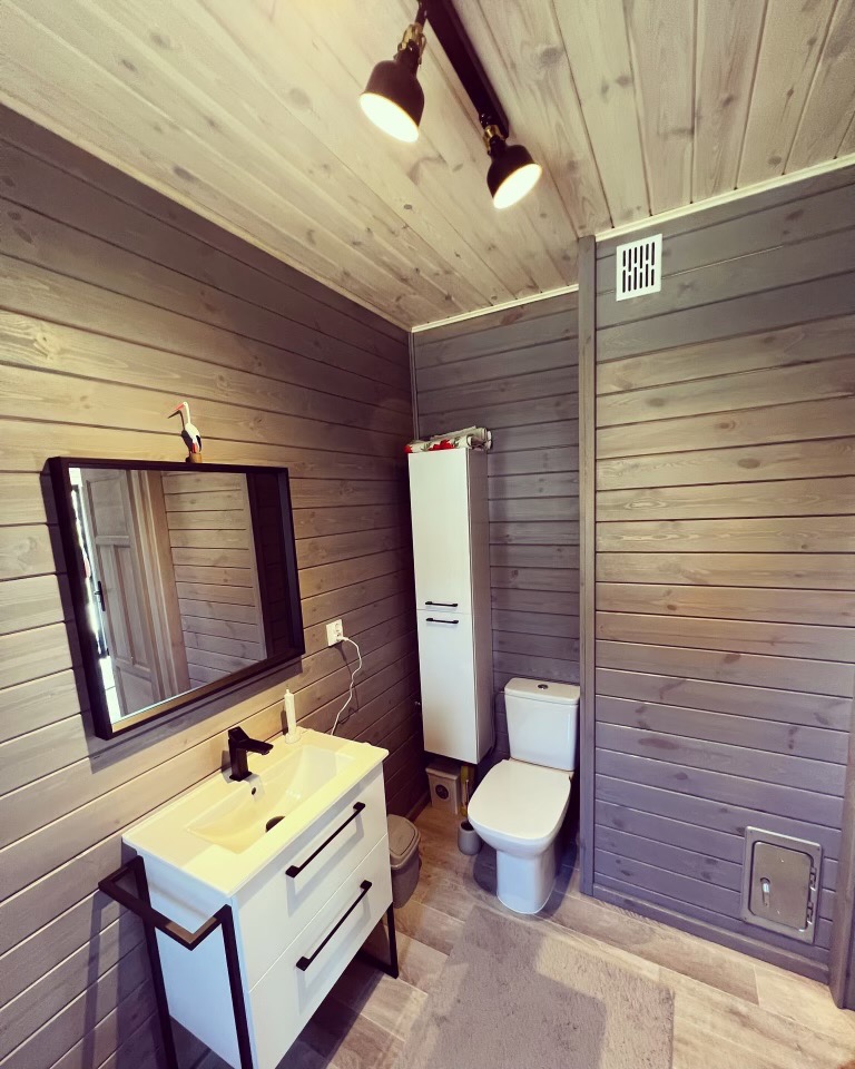 Nowoczesna łazienka z drewnianymi panelami ściennymi w odcieniach szarości, biała szafka z umywalką i czarną armaturą, toaleta oraz biała szafka wisząca. Na umywalce stoi figurka bociana.