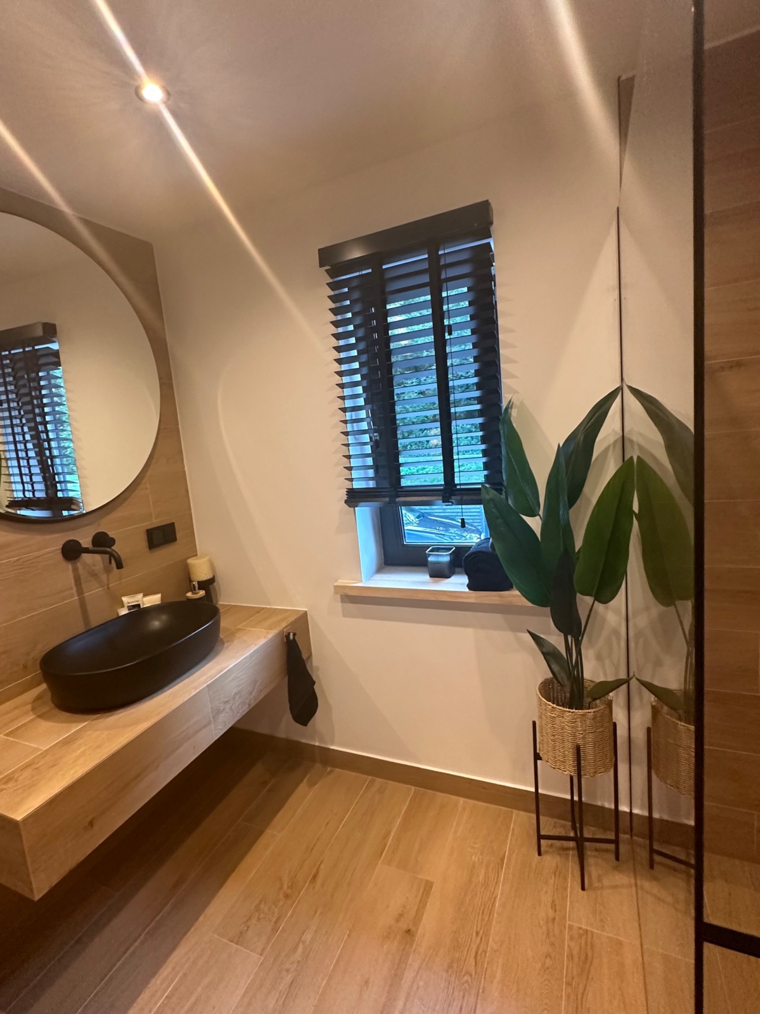 Nowoczesna łazienka z drewnopodobnymi płytkami, okrągłym lustrem, czarną umywalką nablatową i roletami w oknie, uzupełniona rośliną doniczkową w plecionym koszu.