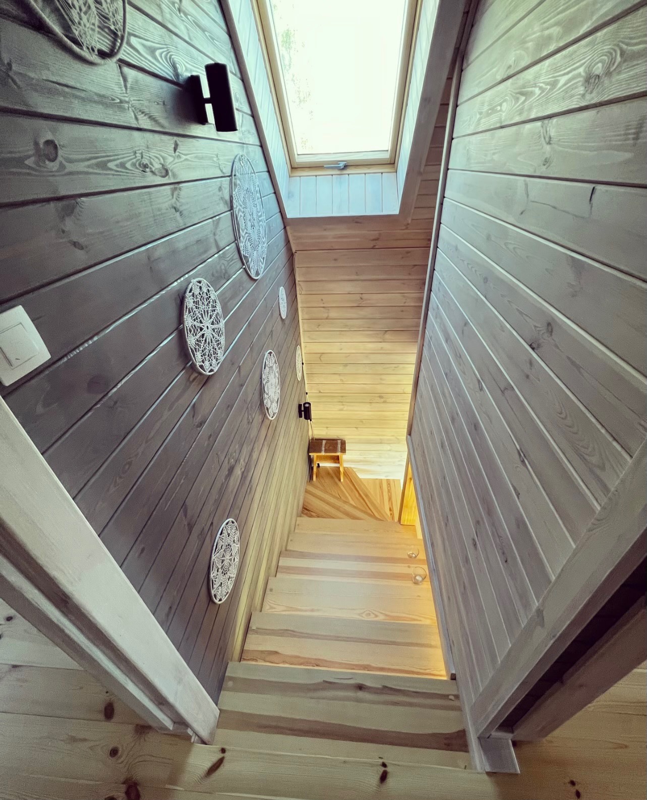 Drewniane schody z oknem dachowym, ściany wykończone boazerią w dwóch odcieniach szarości, ozdobione ażurowymi dekoracjami ściennymi.