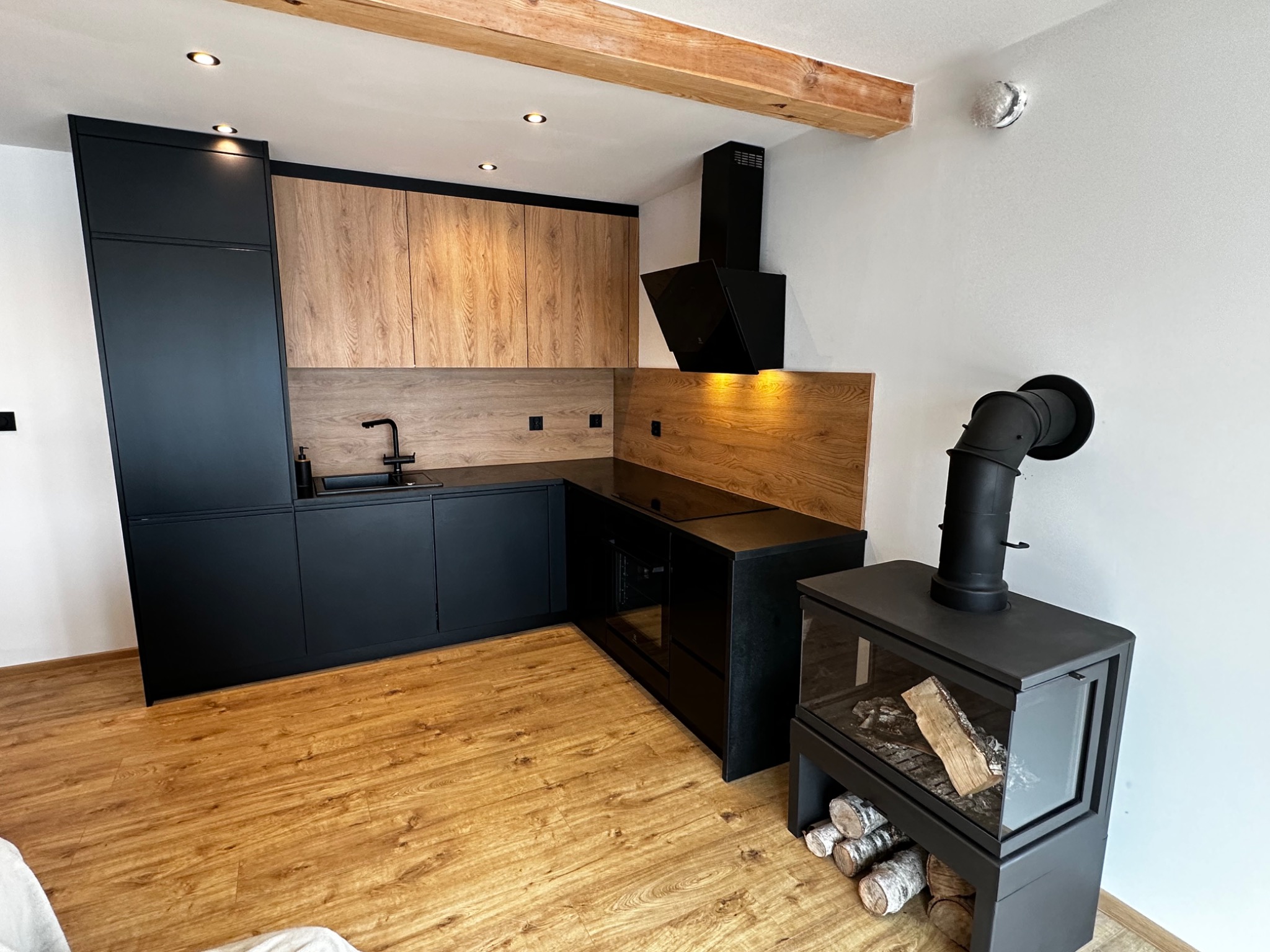 Nowoczesna kuchnia z czarnymi matowymi szafkami i drewnianymi akcentami, oświetleniem punktowym i kominkiem z zapasem drewna, parkiet w odcieniach brązu.