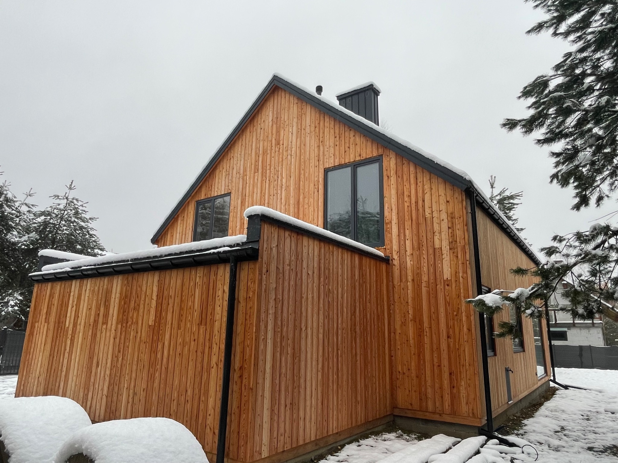 Nowoczesny dom z drewnianą elewacją w zimowej scenerii, widoczne śnieżne czapy na dachu i w ogrodzie, pionowe deski elewacyjne o ciepłym odcieniu drewna, czarne obramowania okien i rynny.