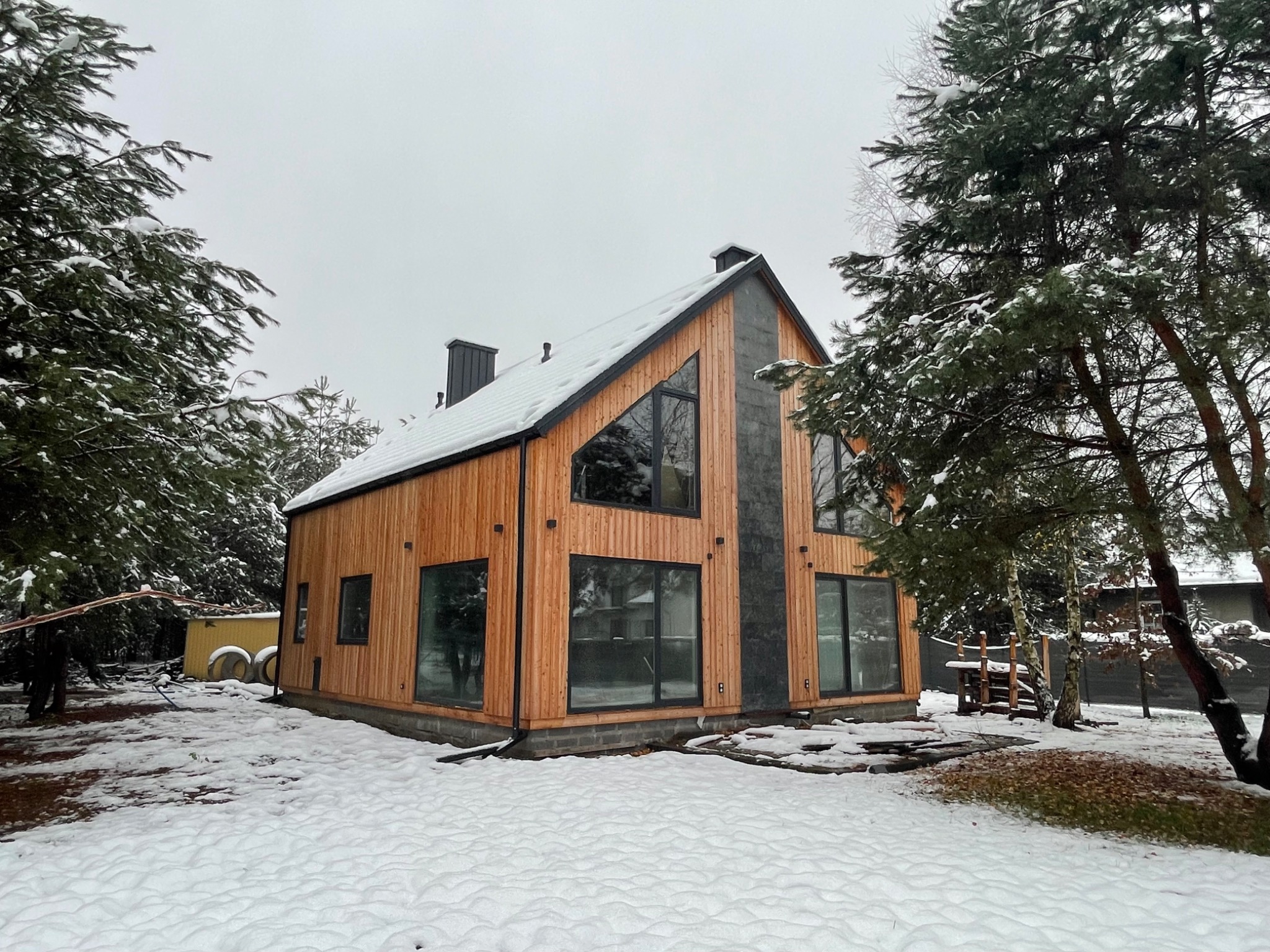 Nowoczesny dom z drewnianą elewacją i dużymi oknami, częściowo pokryty śniegiem, otoczony drzewami w zimowej scenerii.