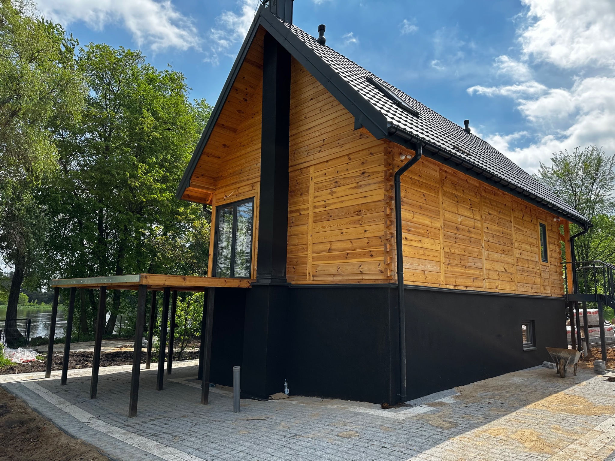 Nowoczesny dom z drewnianą elewacją i czarnym fundamentem, widok z zewnątrz na tle zieleni i błękitnego nieba z chmurami.