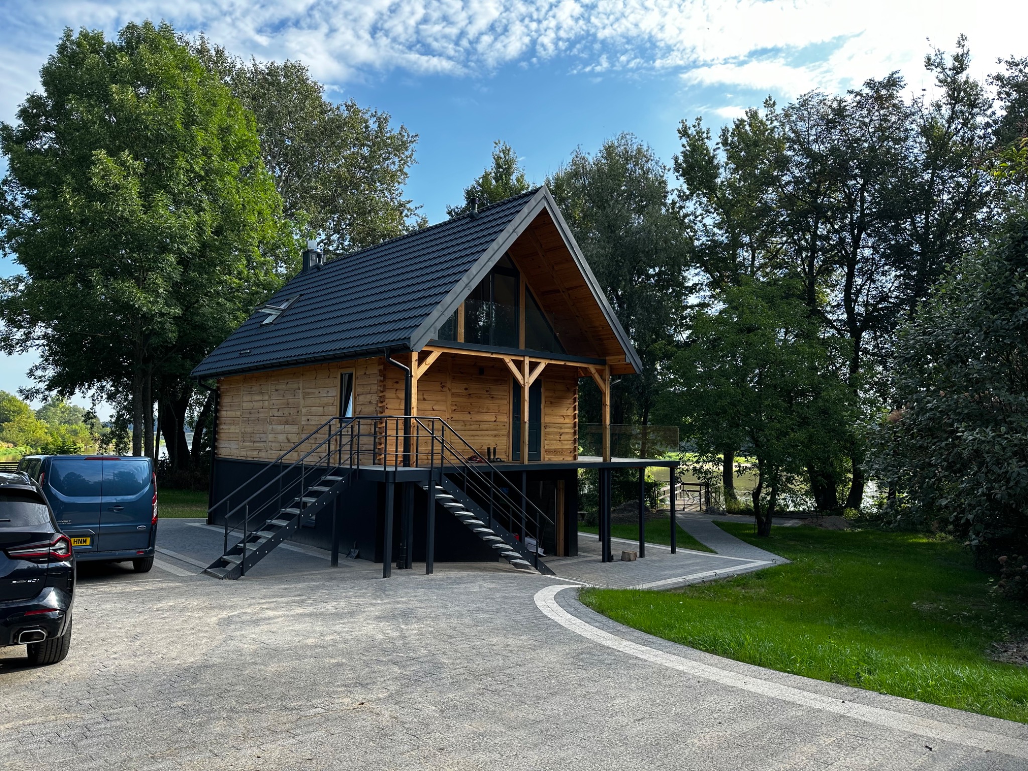 Nowoczesny, drewniany dom na podwyższeniu z czarnymi schodami i dachem z dachówki, otoczony zielenią i samochodami na podjeździe.