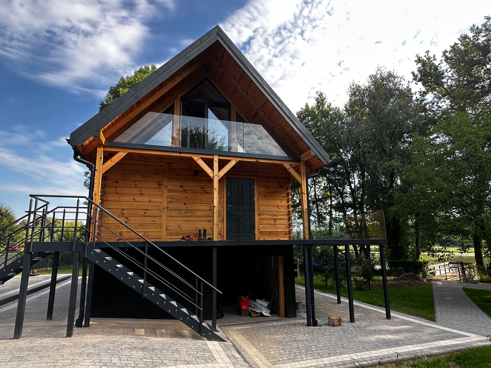Drewniany dom z czarnymi elementami konstrukcyjnymi i szklaną balustradą balkonu, widoczny z zewnątrz, na podwyższeniu, z czarnymi schodami prowadzącymi na taras, otoczony zielenią.
