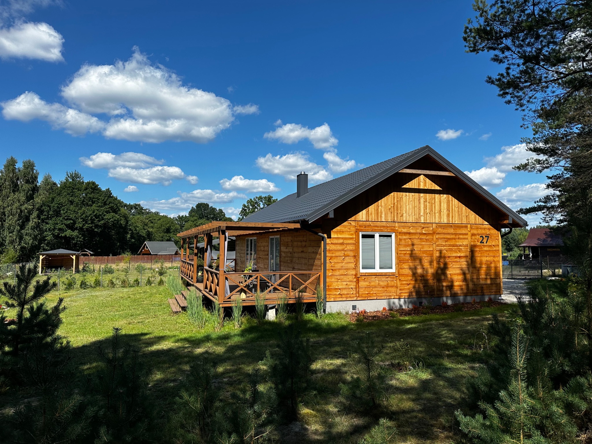 Drewniany dom jednorodzinny z tarasem, numer 27 na ścianie, otoczony zielenią i drzewami, na tle błękitnego nieba z białymi chmurami.