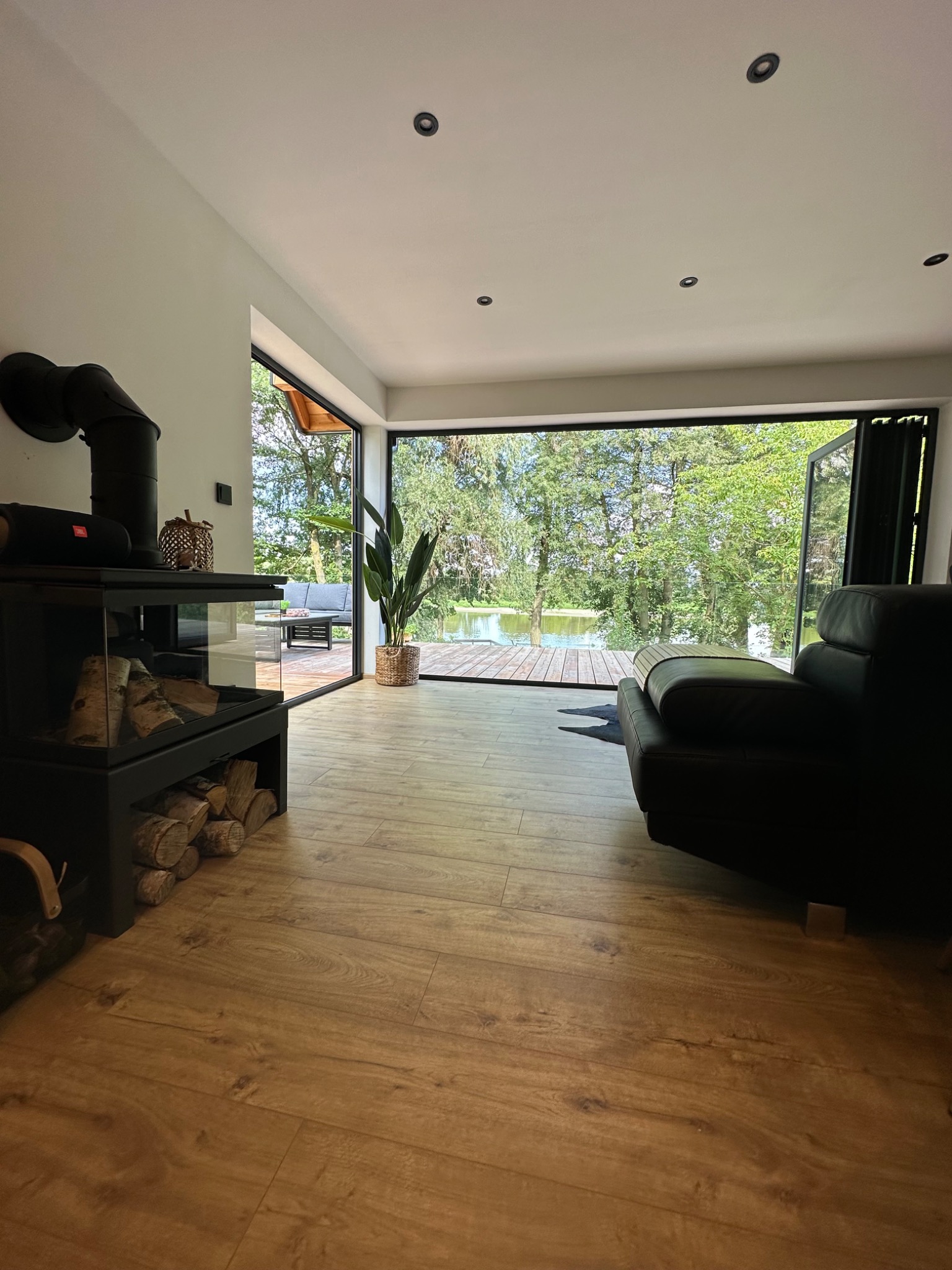 Nowoczesny salon z kominkiem i widokiem na jezioro przez duże okno, drewniana podłoga, czarna sofa i czarny kominek z zapasem drewna.