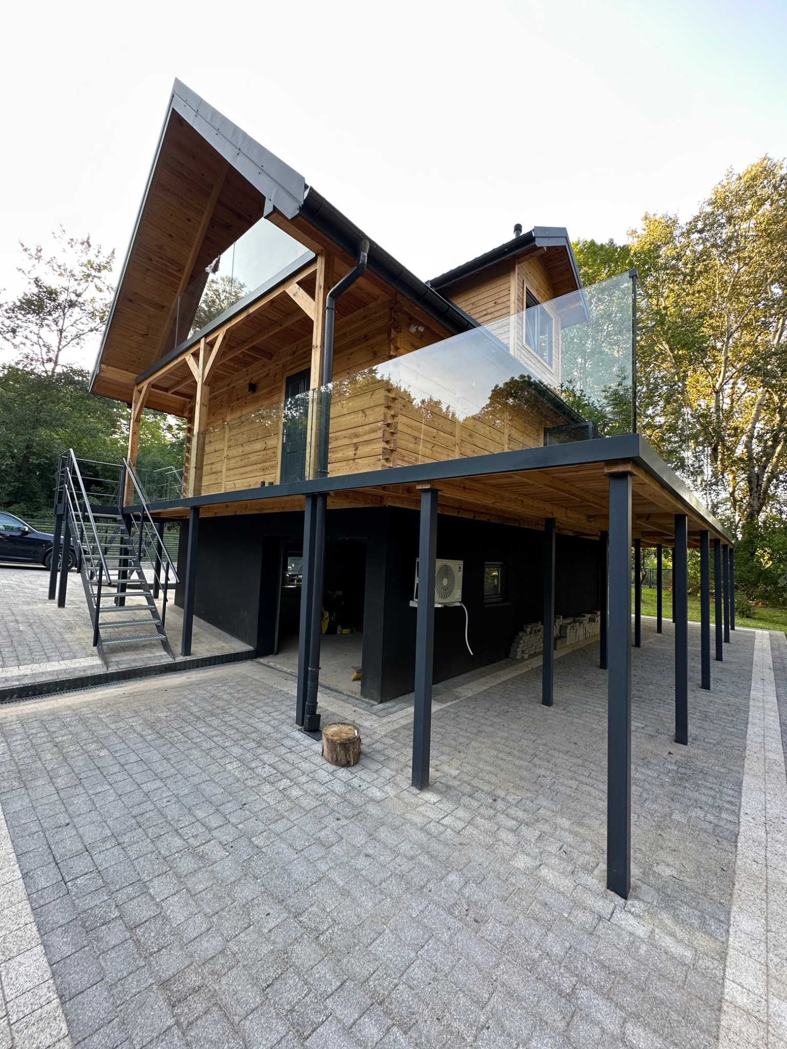 Nowoczesny dom z drewnianą elewacją, szklanymi balustradami balkonu i czarnymi filarami, otoczony brukowanym podjazdem i zielenią.