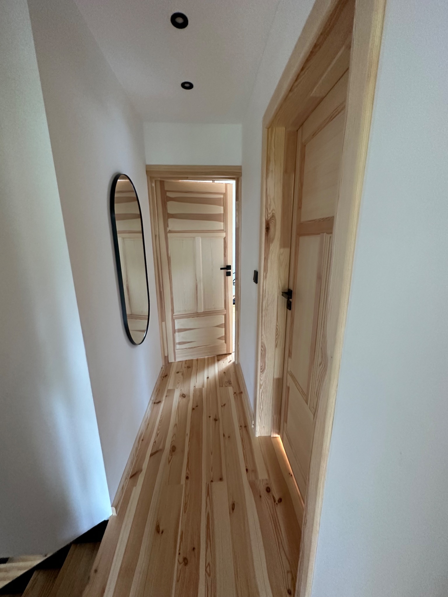 Wąski korytarz z drewnianą podłogą, białymi ścianami, owalnym lustrem i dwiema drewnianymi ościeżnicami z widocznymi drzwiami.