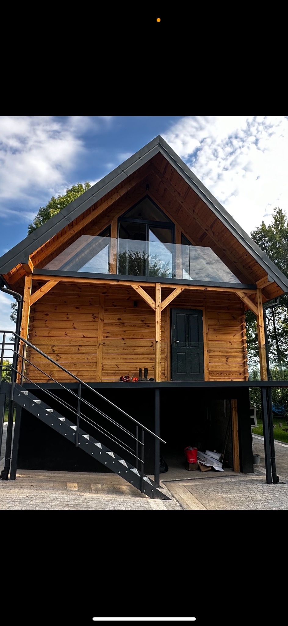 Nowoczesny, drewniany dom z czarnymi elementami konstrukcyjnymi i szklaną balustradą balkonu, widoczny z zewnątrz, z metalowymi schodami wejściowymi i częściowo otwartym parterem.