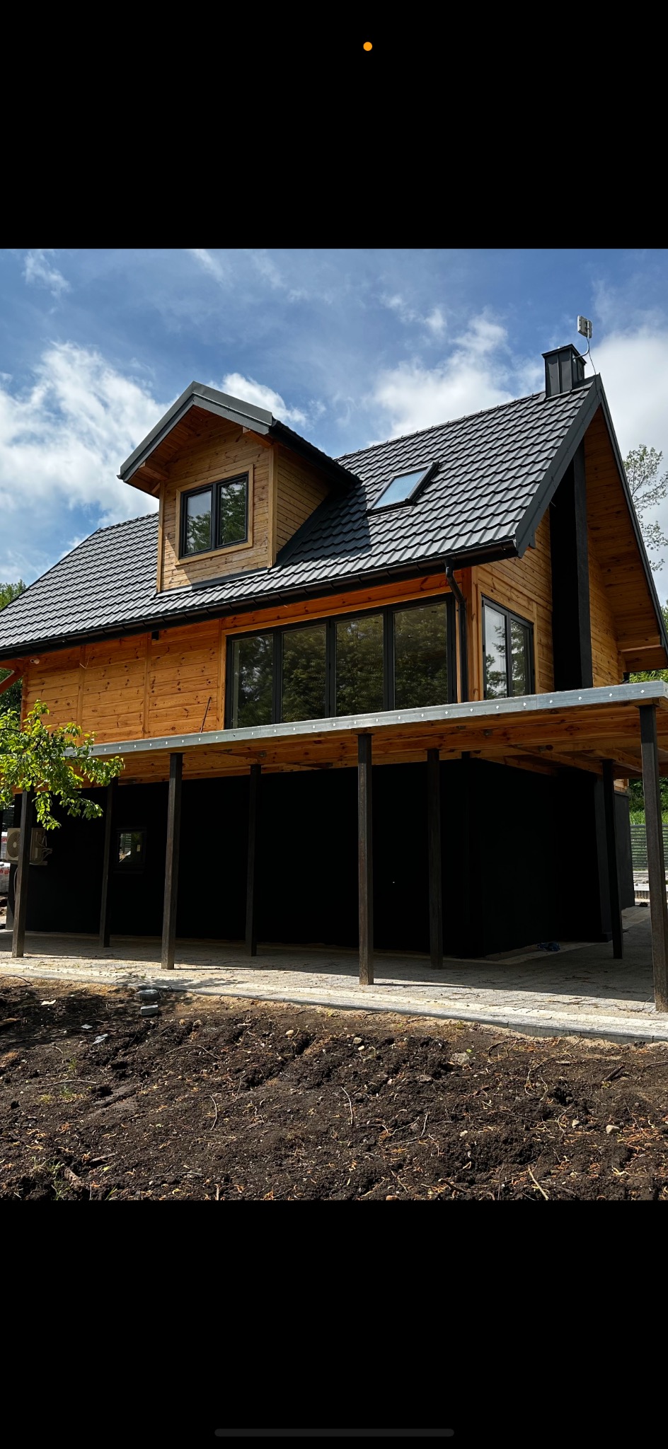 Drewniany dom z czarnymi elementami wykończeniowymi, widoczne okna z czarnymi ramami, dach z ciemnej dachówki i odsłonięta ziemia przed budynkiem.