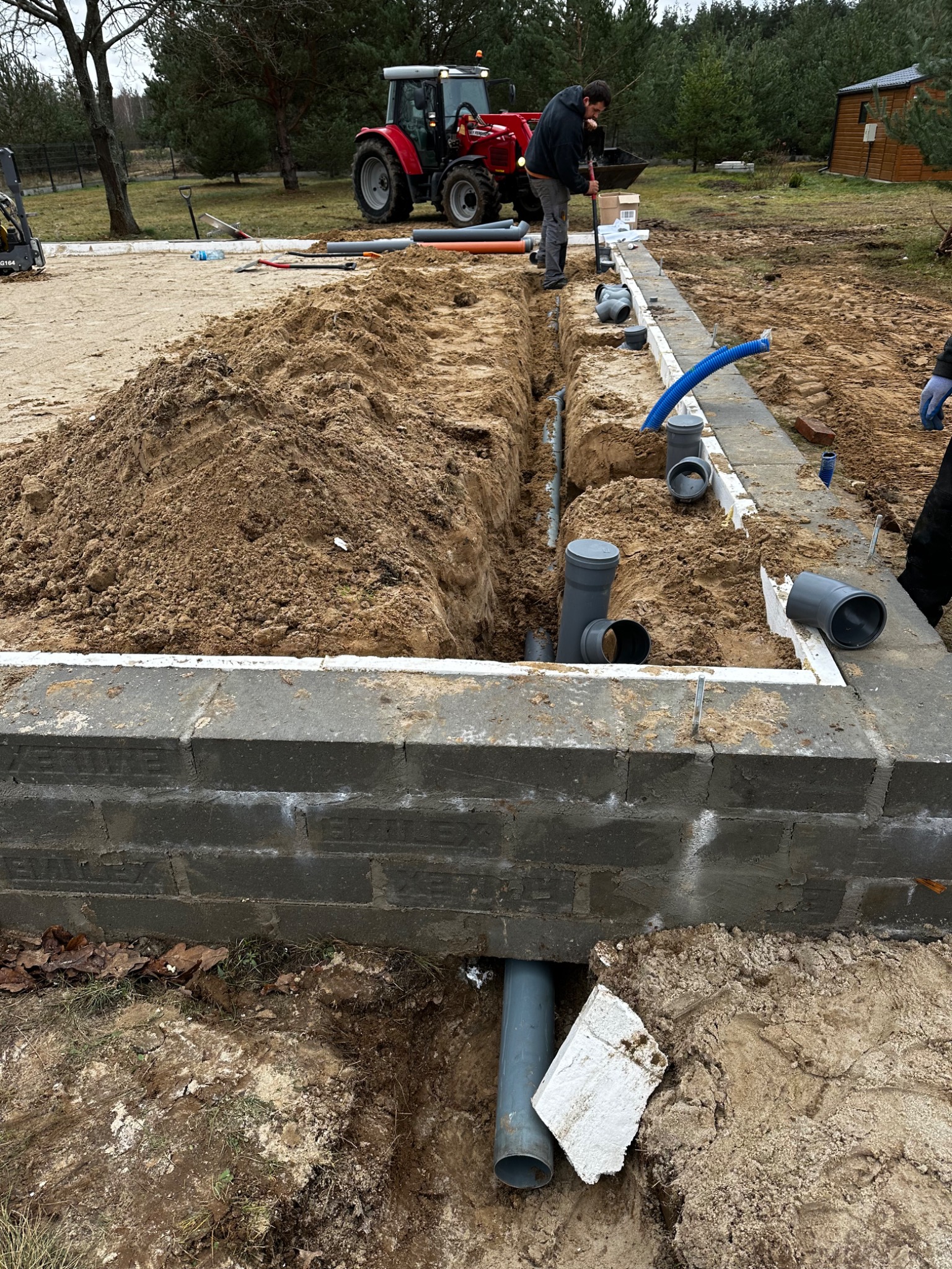 Fundament budynku w trakcie budowy z widocznymi rurami kanalizacyjnymi w wykopie, w tle czerwony traktor i pracownik z łopatą.